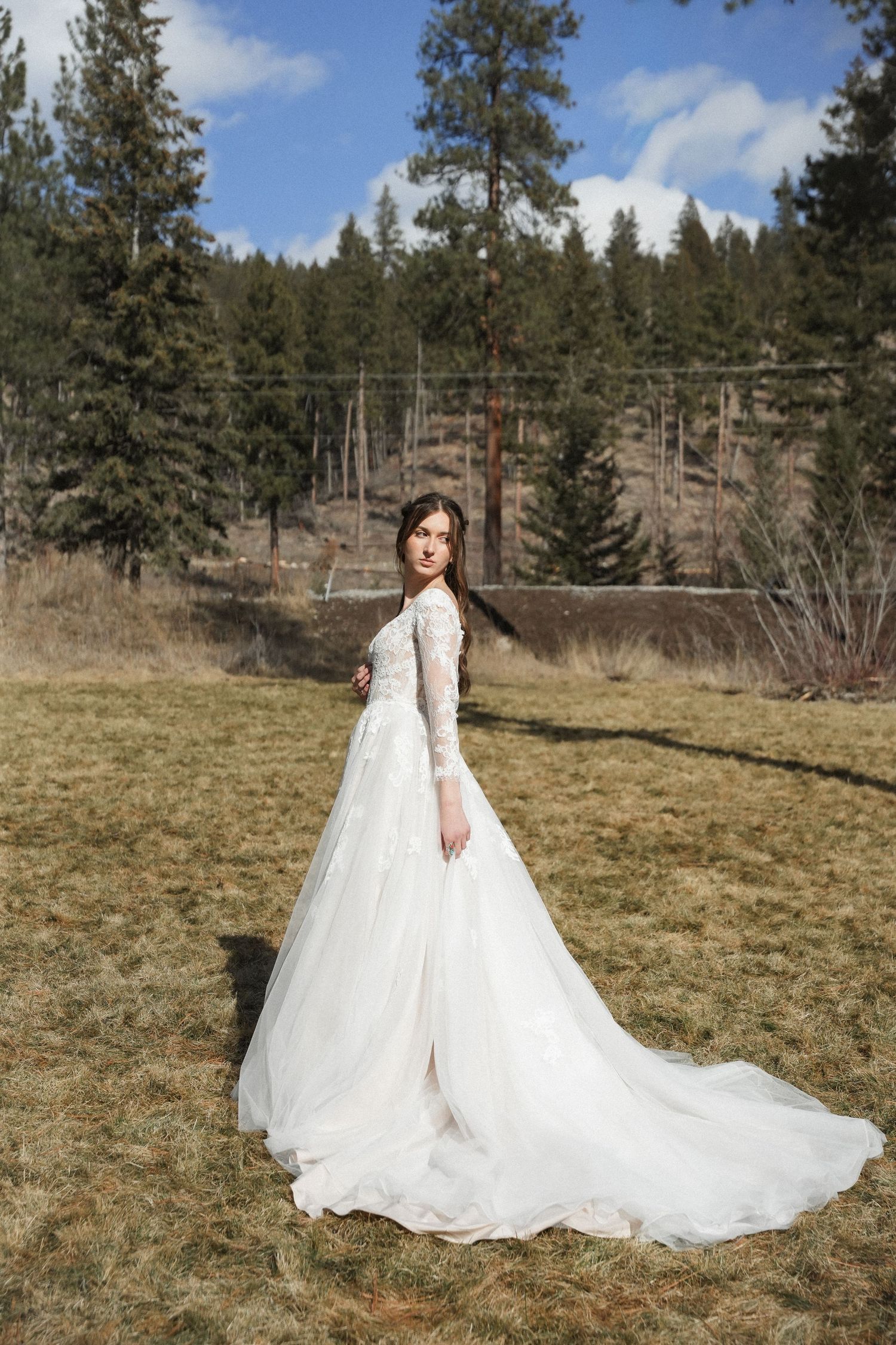 A bride in a flowing white lace wedding dress poses in a rustic outdoor setting with pine trees in the background.