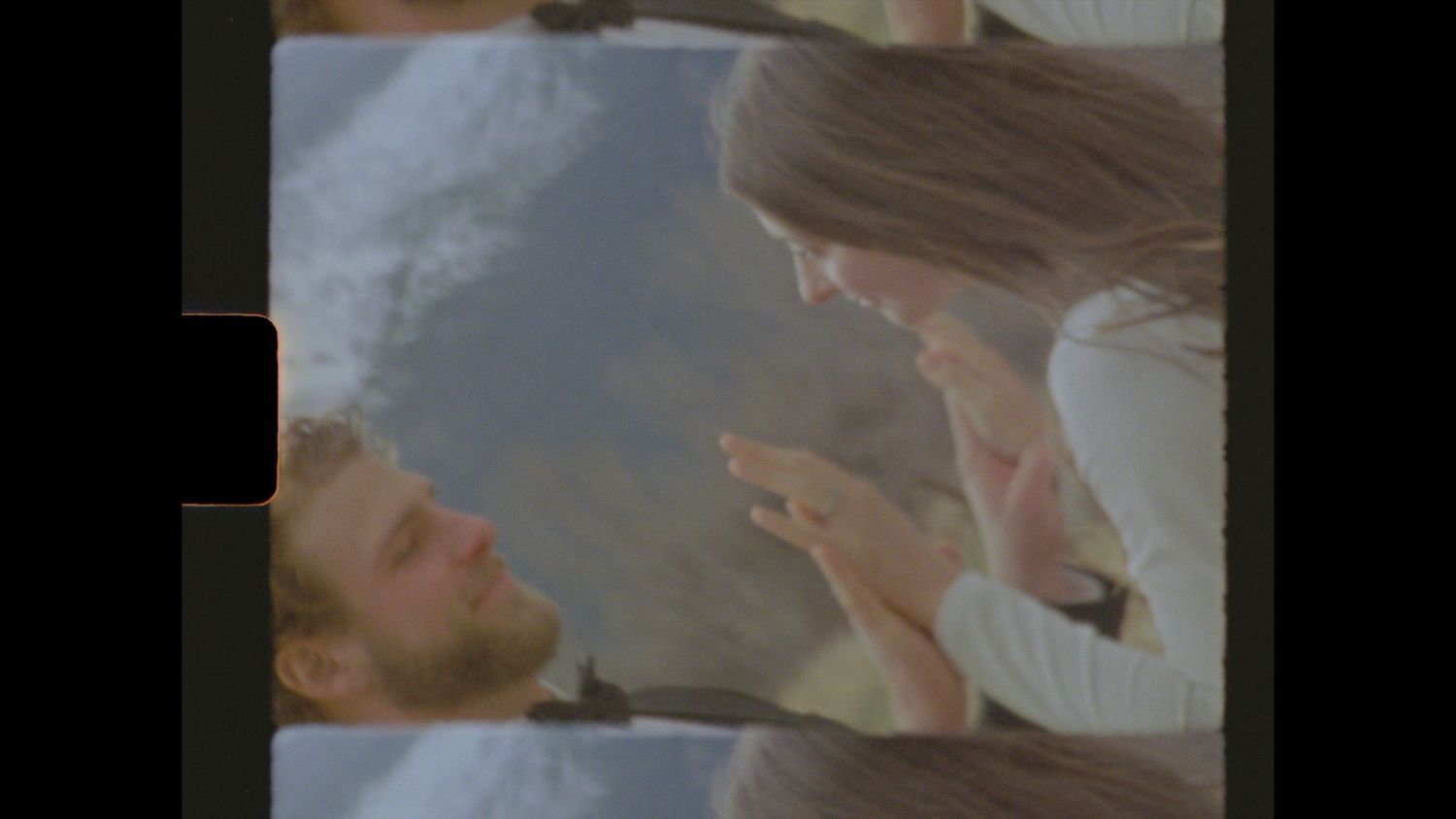 A bearded man lying down looks up at a woman with long hair pointing her finger at him outdoors.