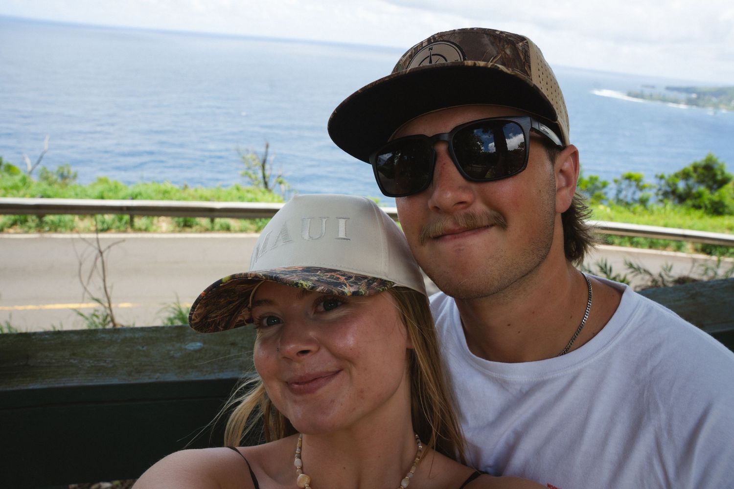 A smiling couple wearing baseball caps poses outdoors with a scenic ocean view and lush greenery in the background.