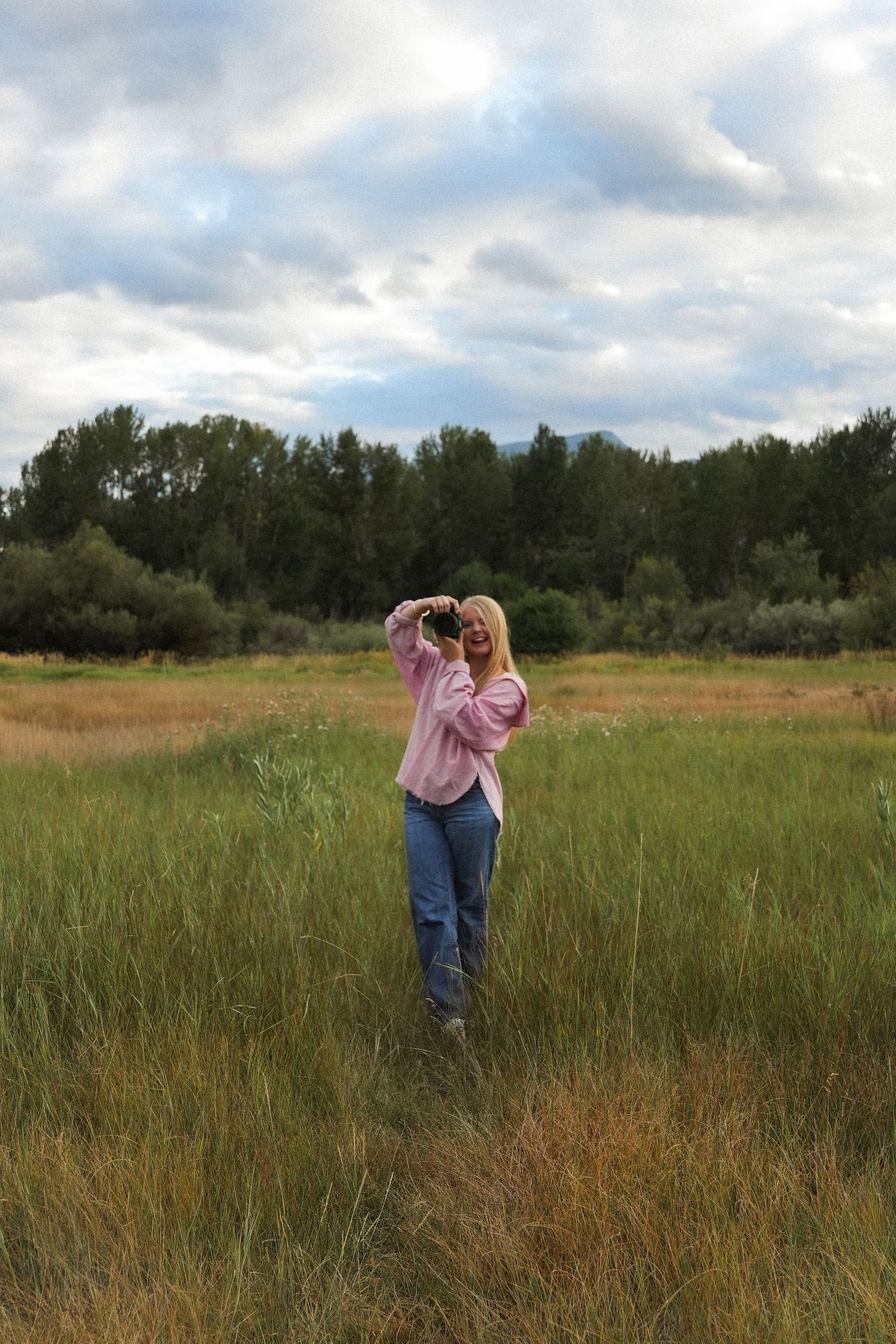 Blonde woman in pink jacket and jeans photographing nature in a lush green meadow under cloudy skies.