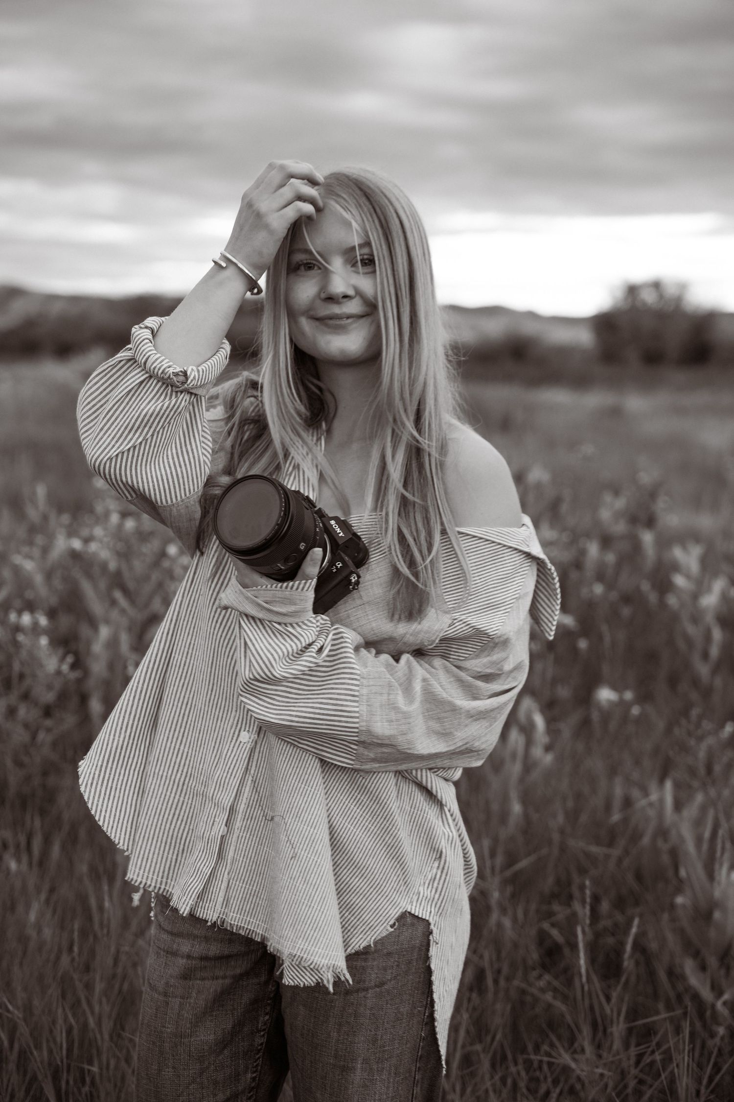Black and white photo of a smiling blonde woman holding a camera in a field, wearing a striped shirt and jeans.