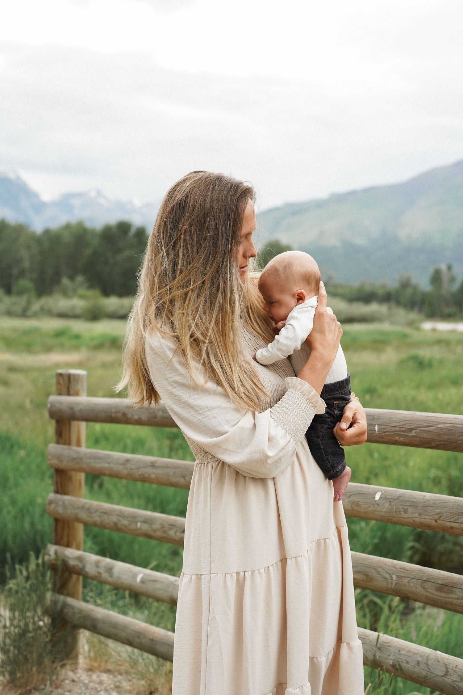A mother in a cream dress holds her baby outdoors near a wooden fence with green meadows and mountains behind her.