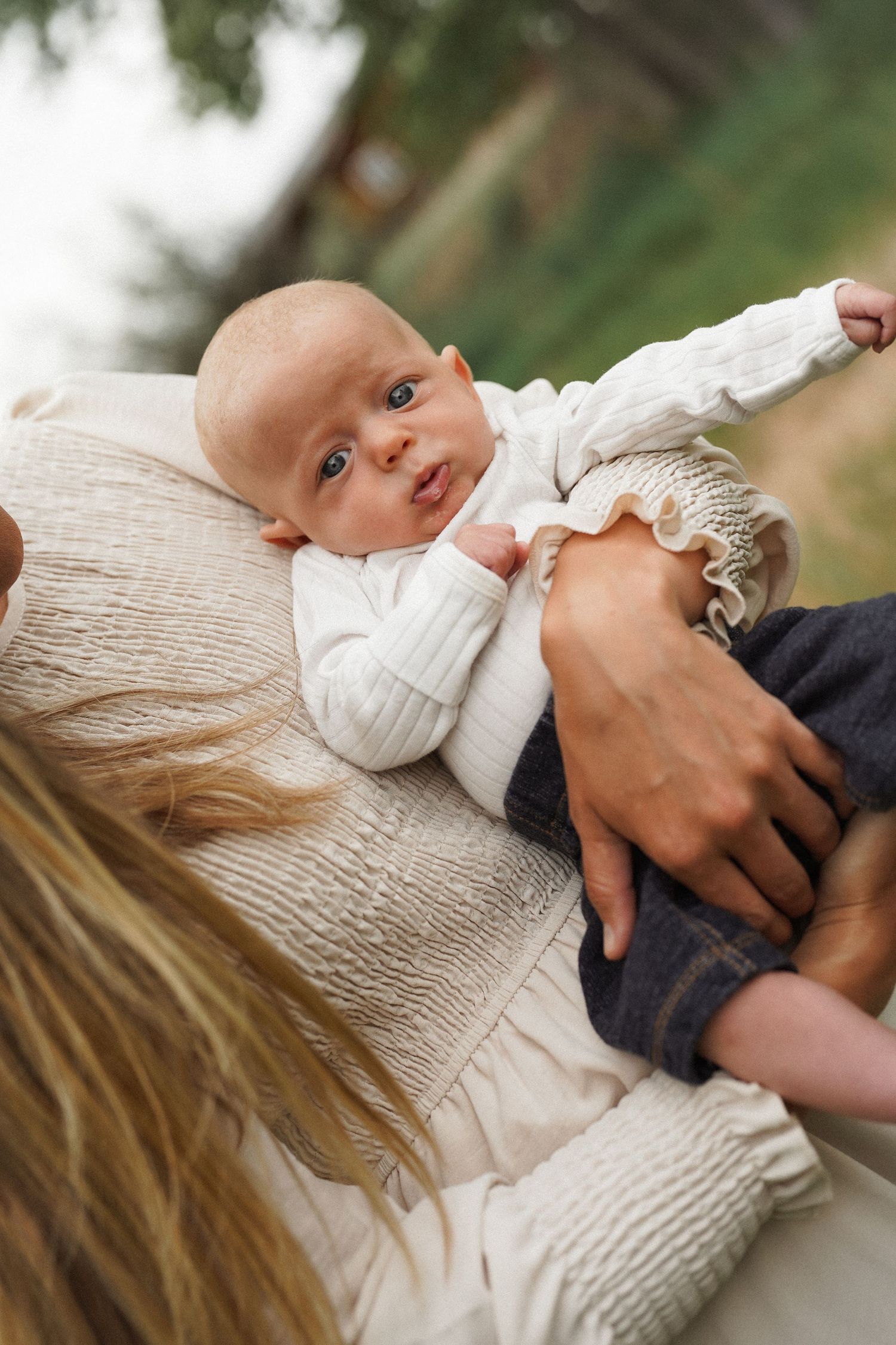 A newborn baby in white outfit resting on mother's shoulder outdoors, looking at camera with curious eyes.