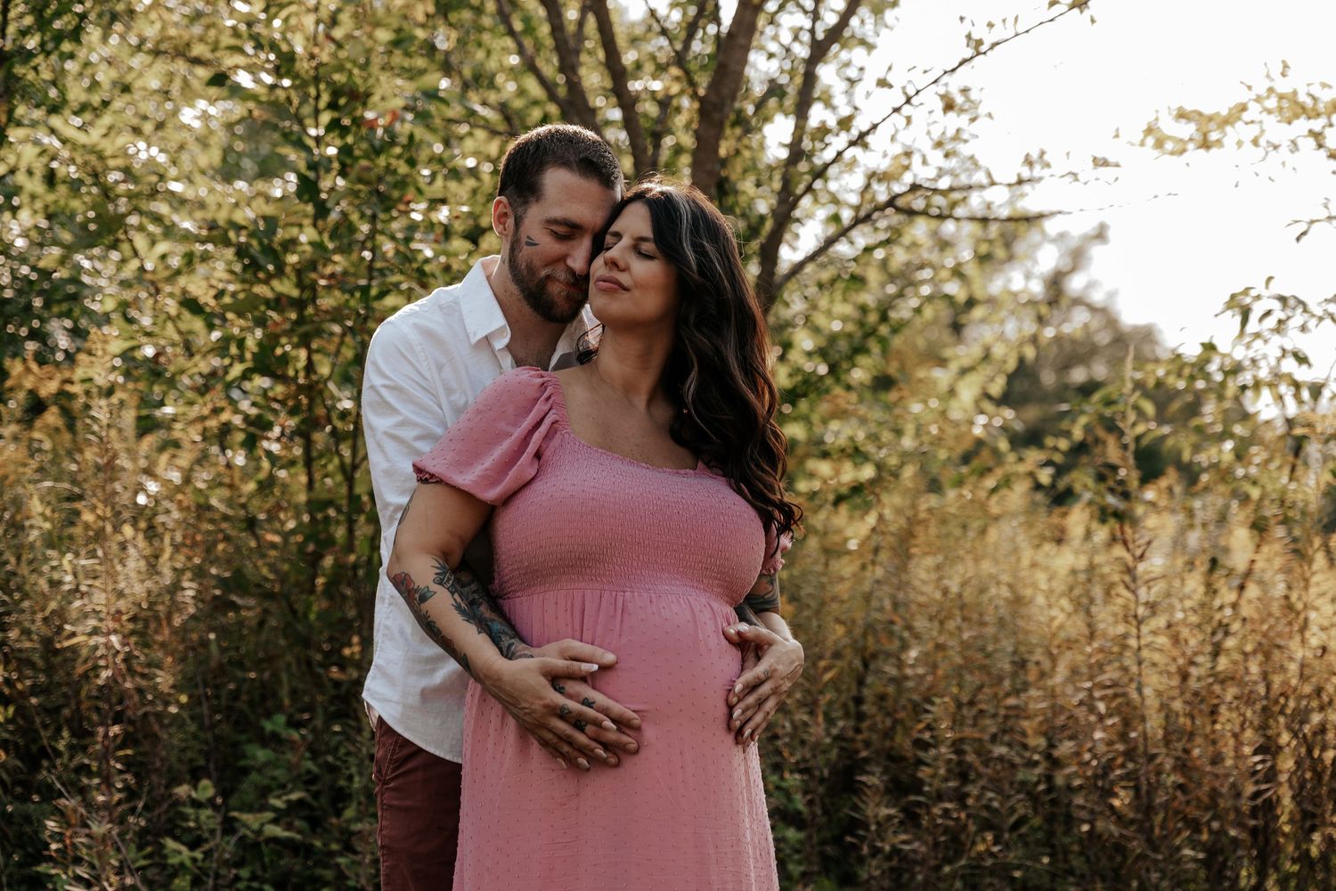 A couple in pink and gray outfits embraces in a golden field during sunset for a romantic photoshoot.