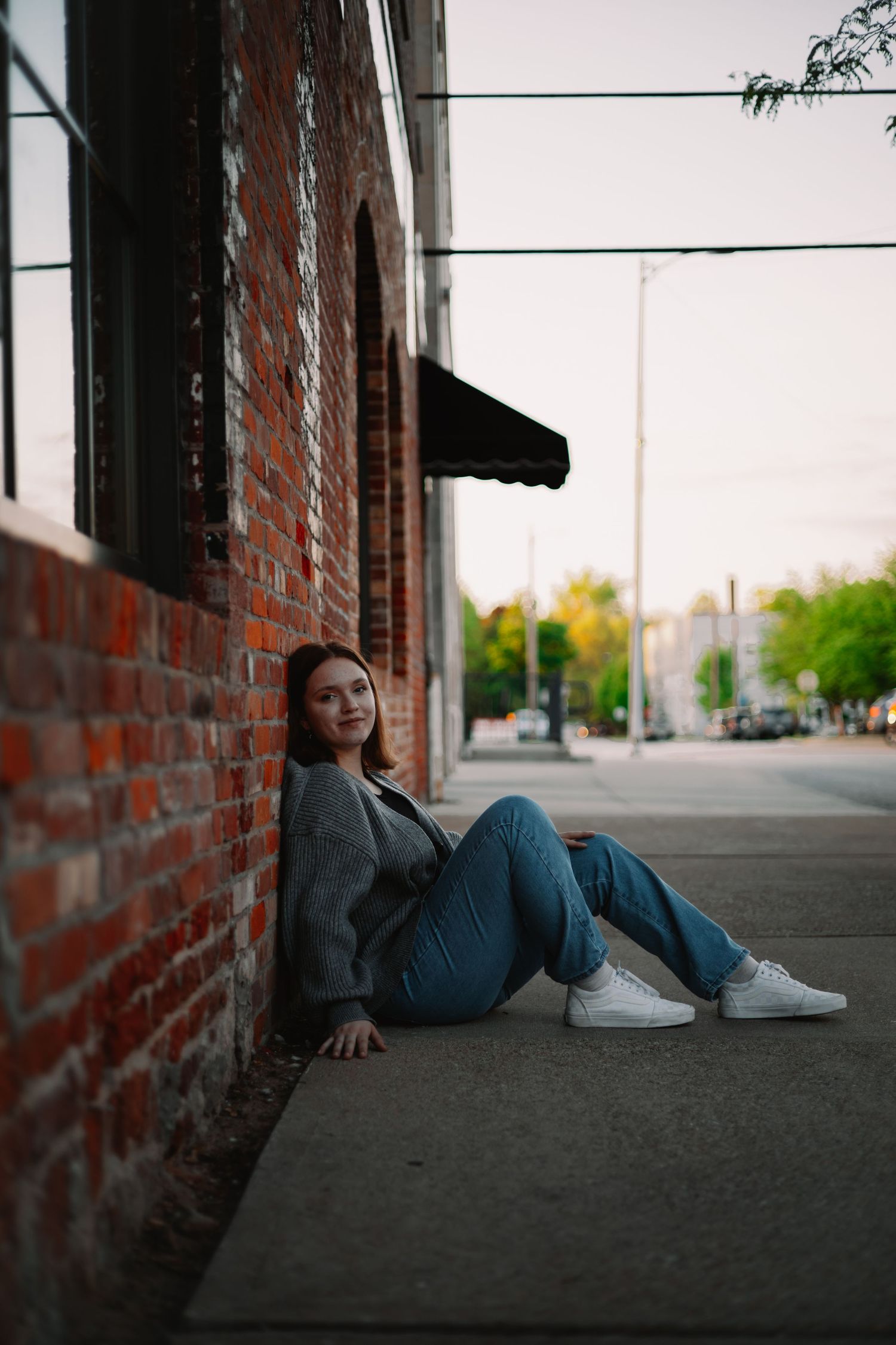 Side view of person reclining on ground against brick wall while wearing casual outfit and sneakers.