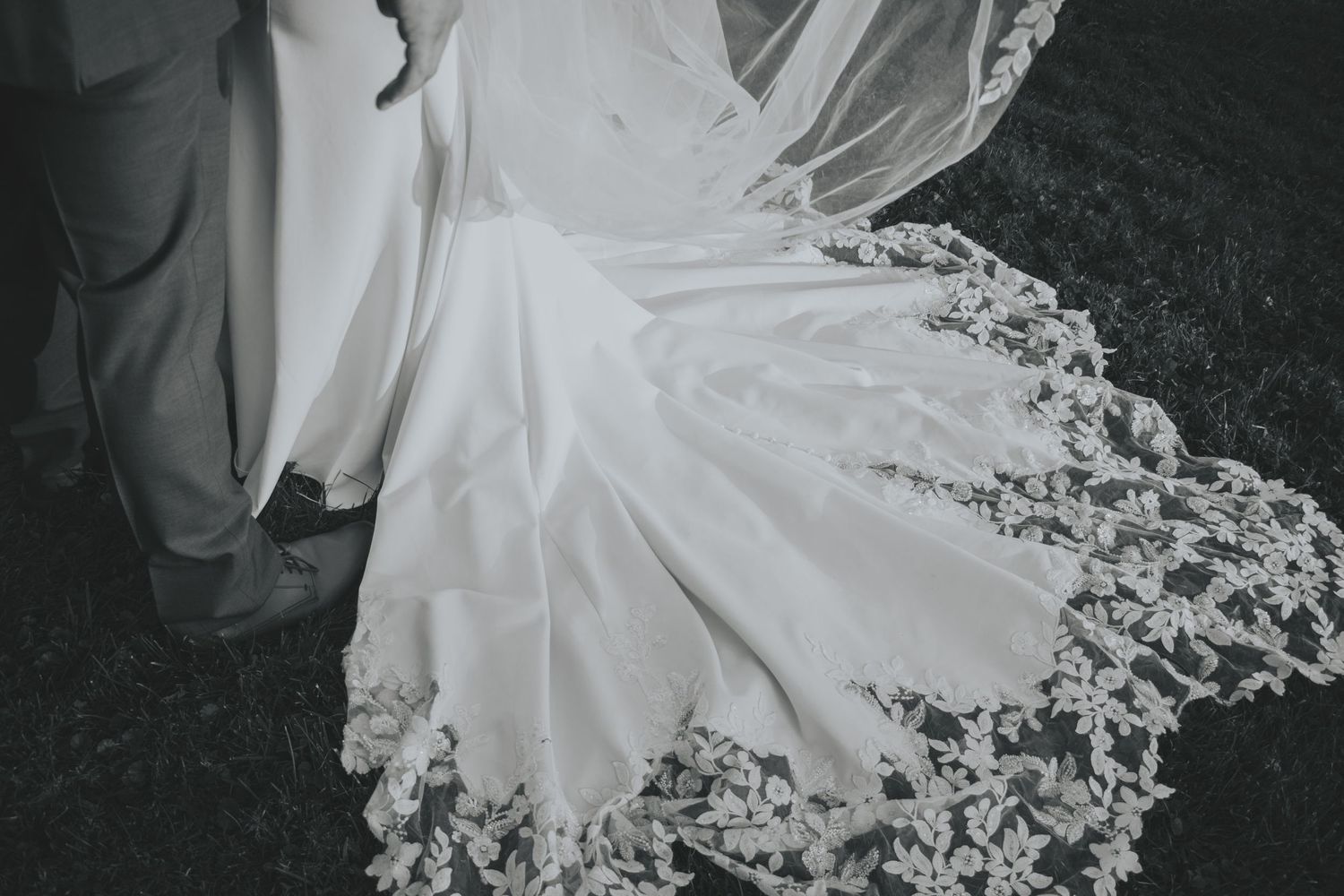 Flowing white wedding dress train with delicate lace details spreads elegantly across dark floor.