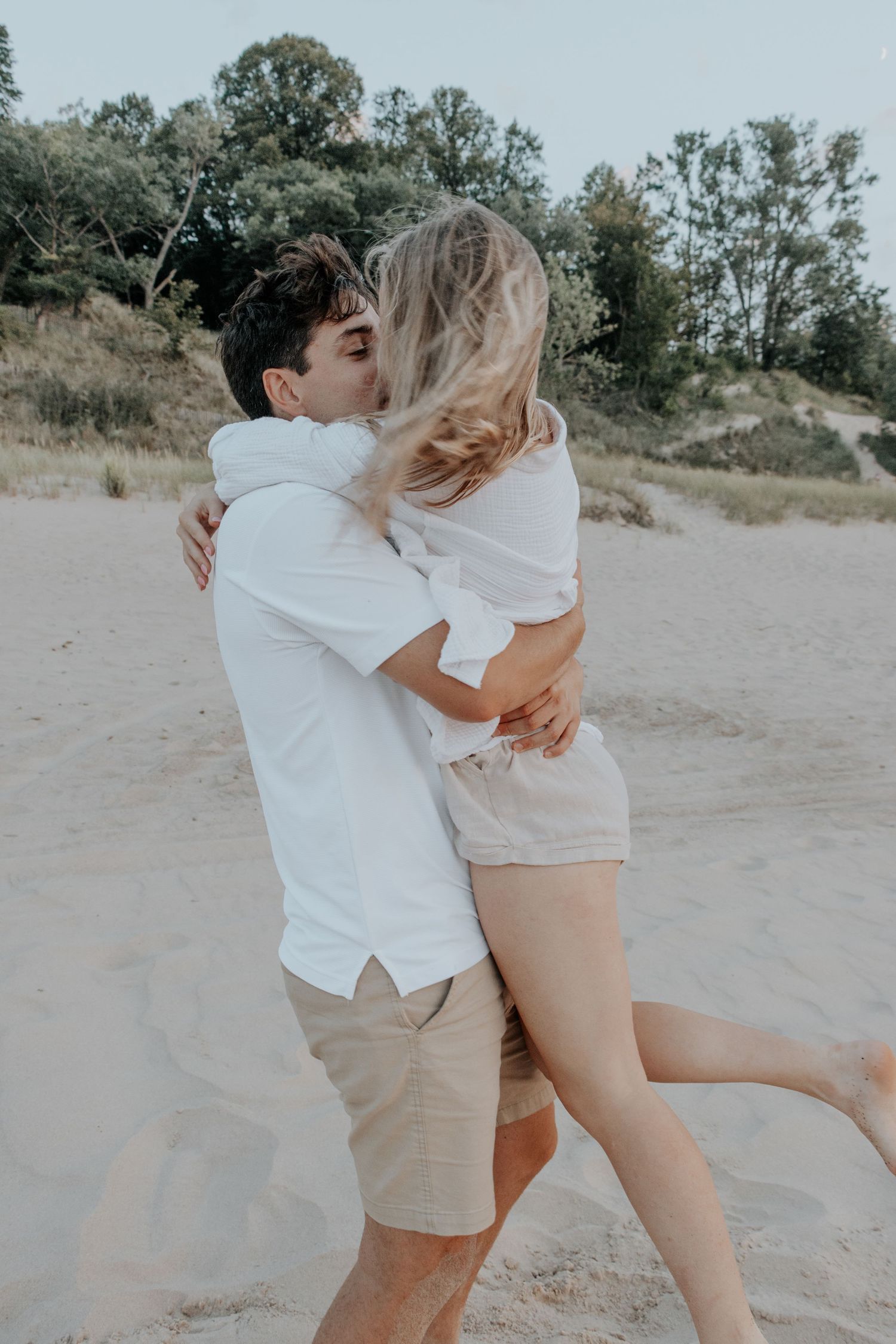 A romantic couple embracing and kissing on a sandy beach surrounded by trees and natural scenery.