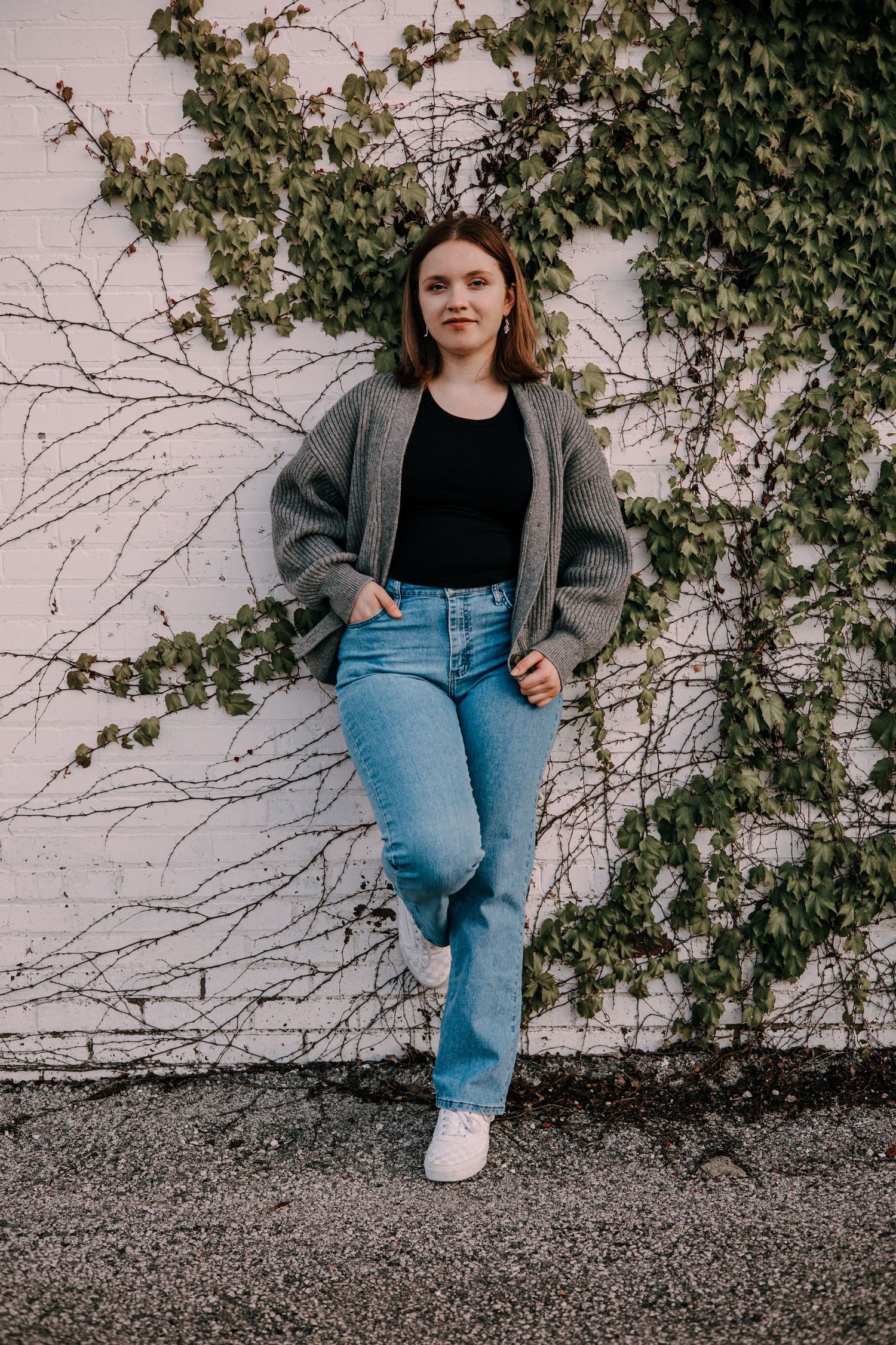 Person wearing gray blazer and blue jeans sits against ivy-covered wall in casual outdoor fashion photoshoot series.