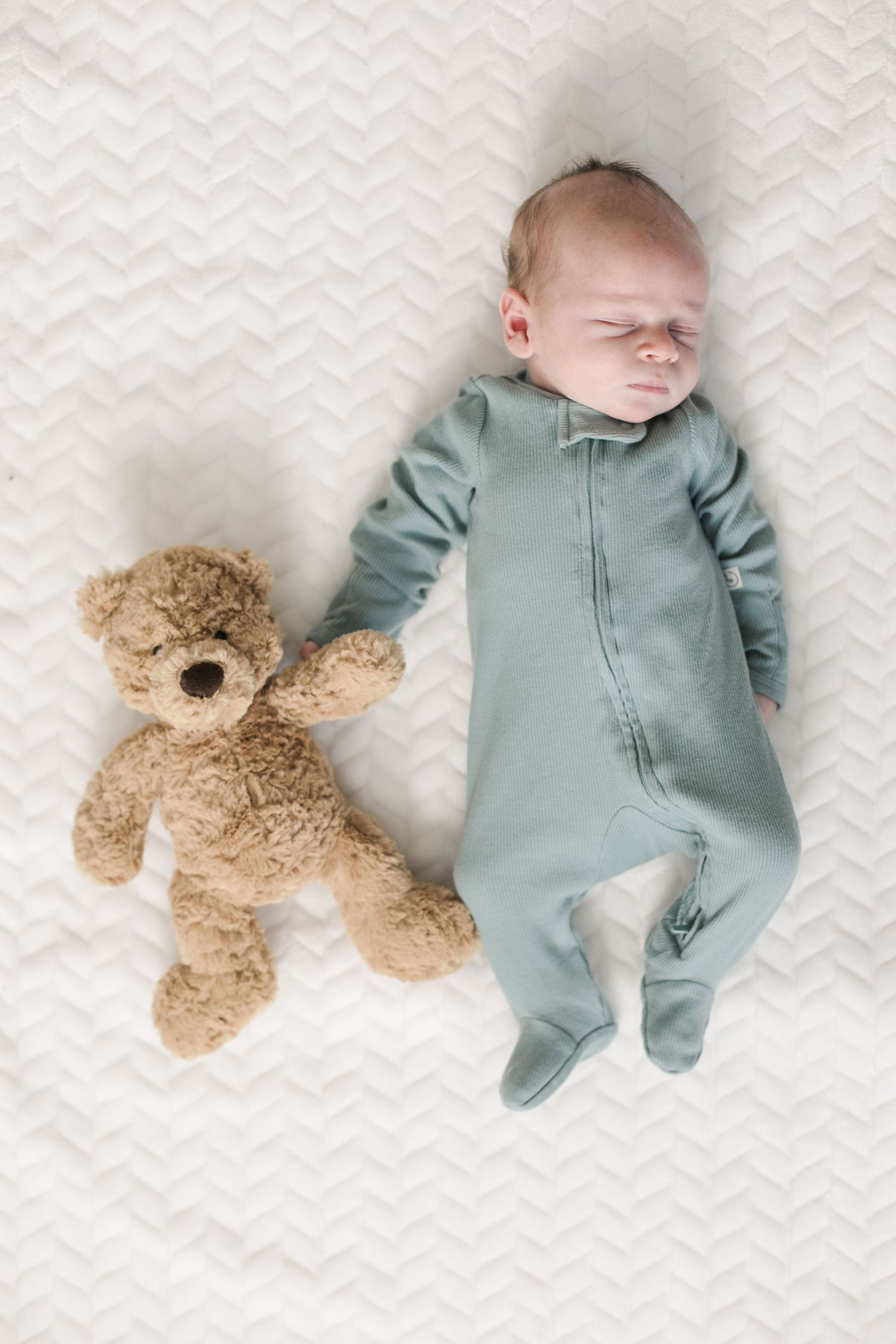newborn baby dressed in blue onesie holding hands with a teddy bear