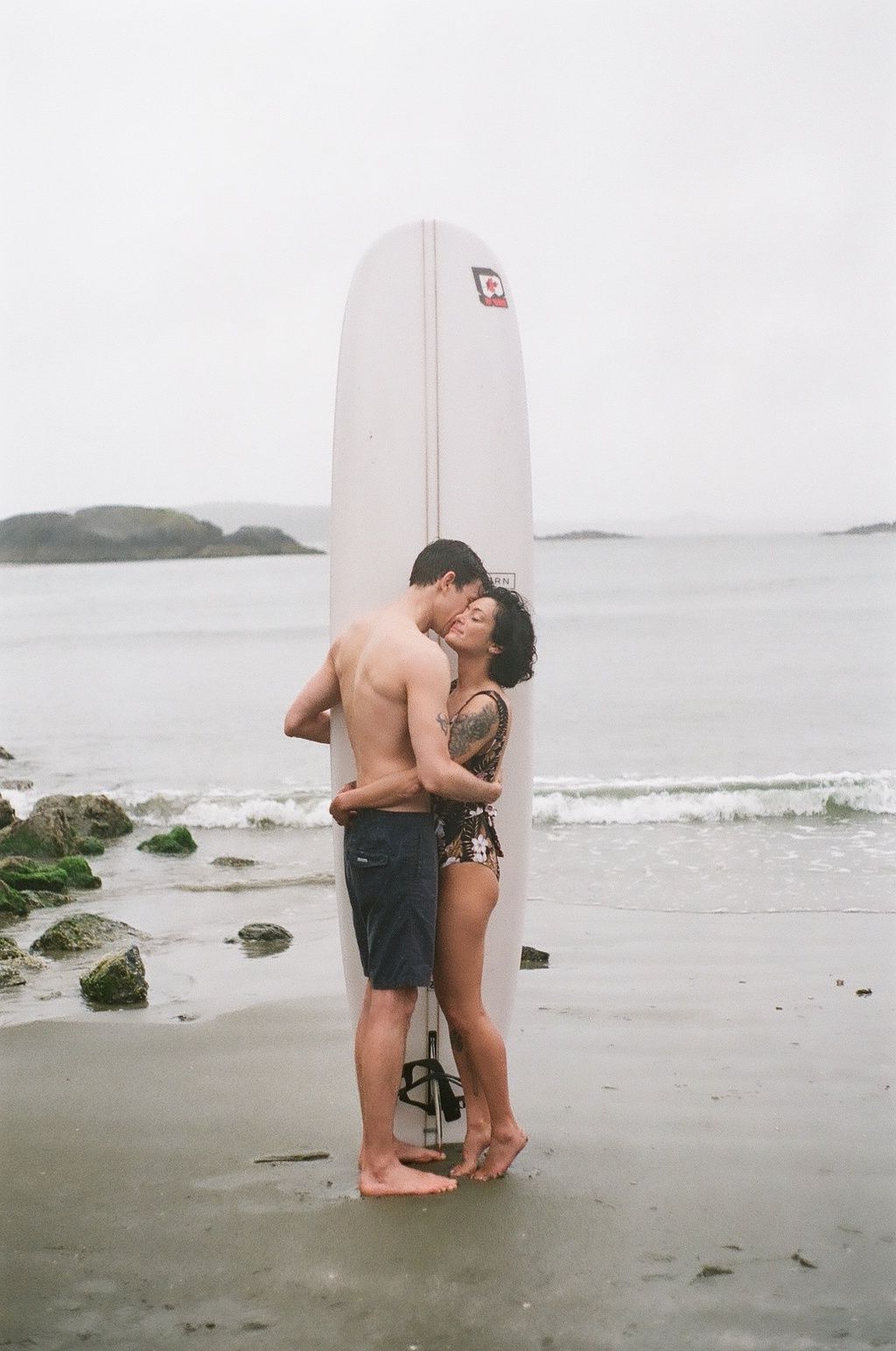 A couple shares an intimate moment on a beach with a surfboard.