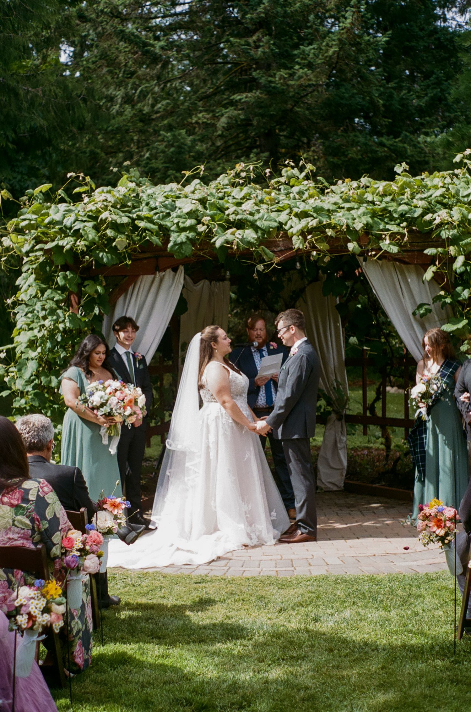 A wedding ceremony takes place under a lush garden arch with white curtains and floral decorations.
