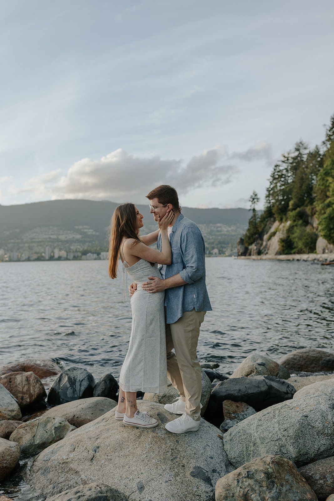 Couple shares intimate moments on rocky shoreline with mountain and lake views.