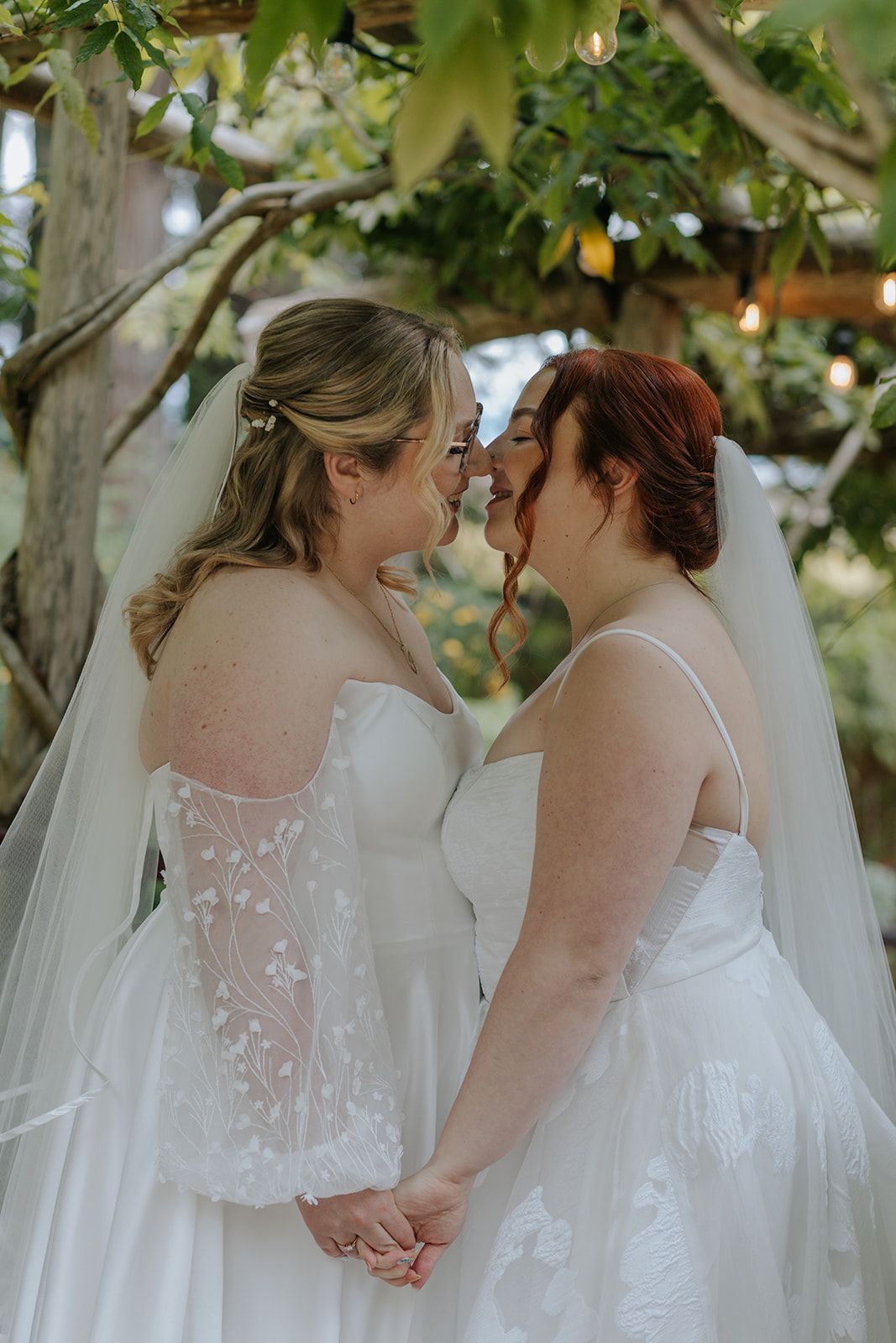 A tender moment between newlyweds in white lace dresses beneath flowering trees.