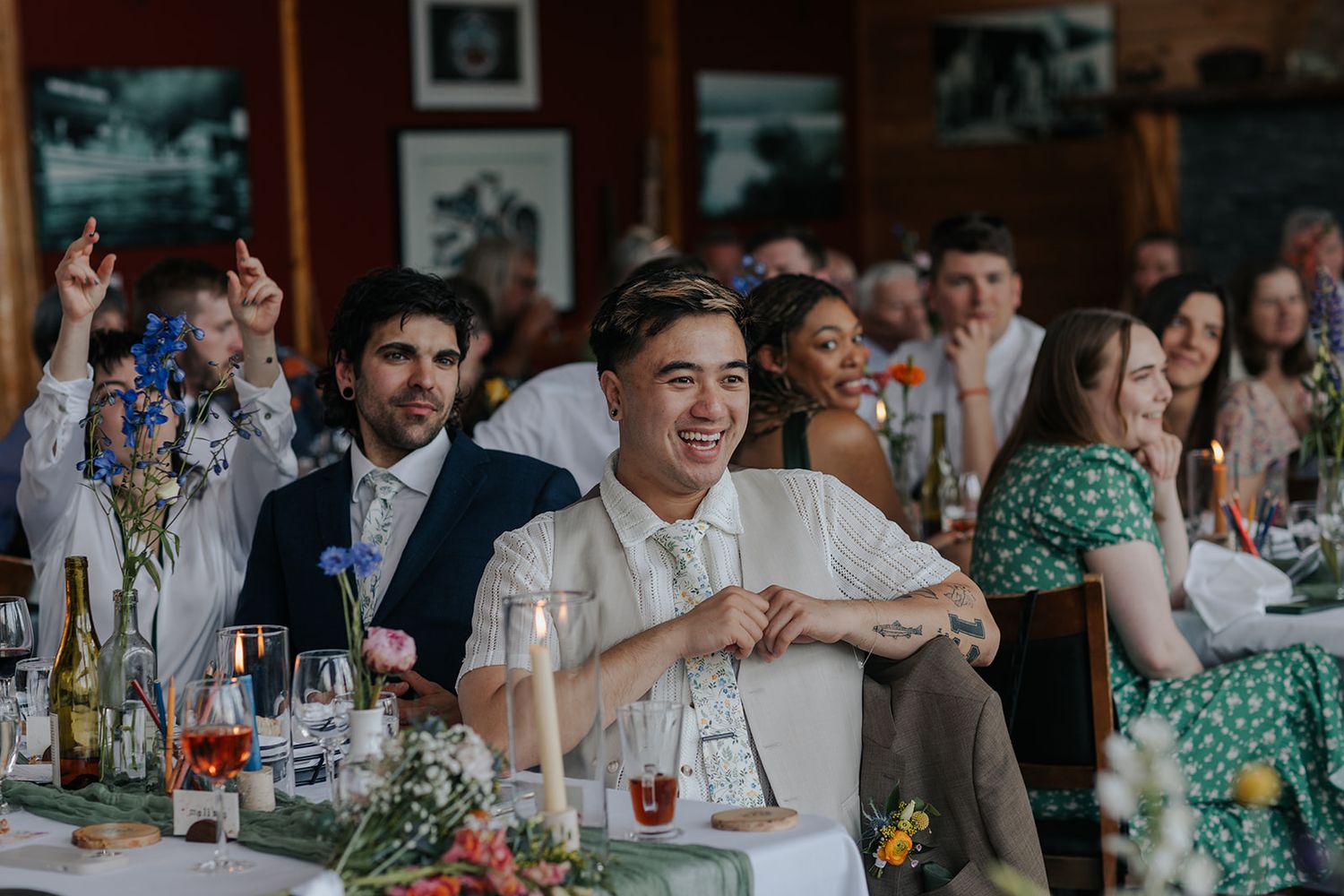 Wedding guests react with laughter during reception speeches at decorated dining table.