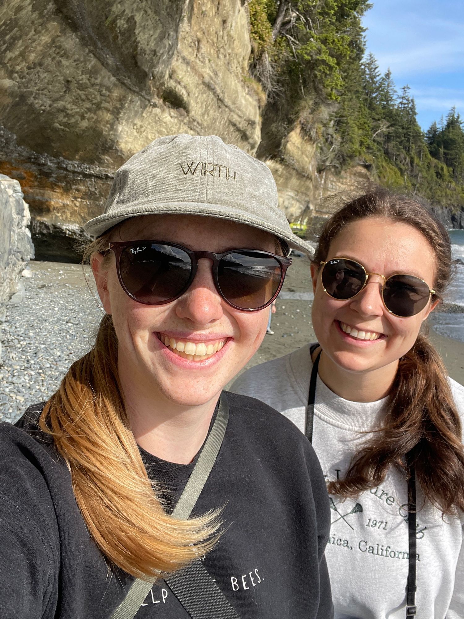 Friends wearing sunglasses and hats taking selfies at rocky beach overlook.