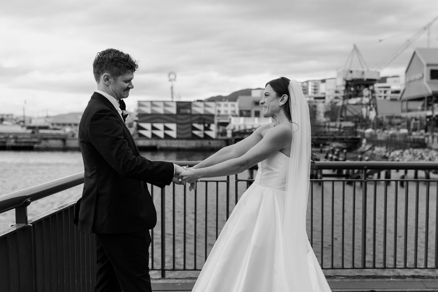 A black and white wedding photo of a couple holding hands on a waterfront deck with buildings in the background.
