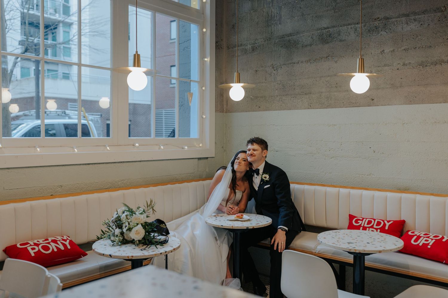 A wedding couple shares an intimate moment on a banquette with hanging pendant lights and floral arrangements.