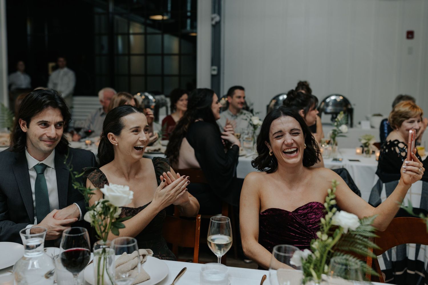 Wedding guests laugh and celebrate together at reception dinner tables decorated with floral centerpieces.