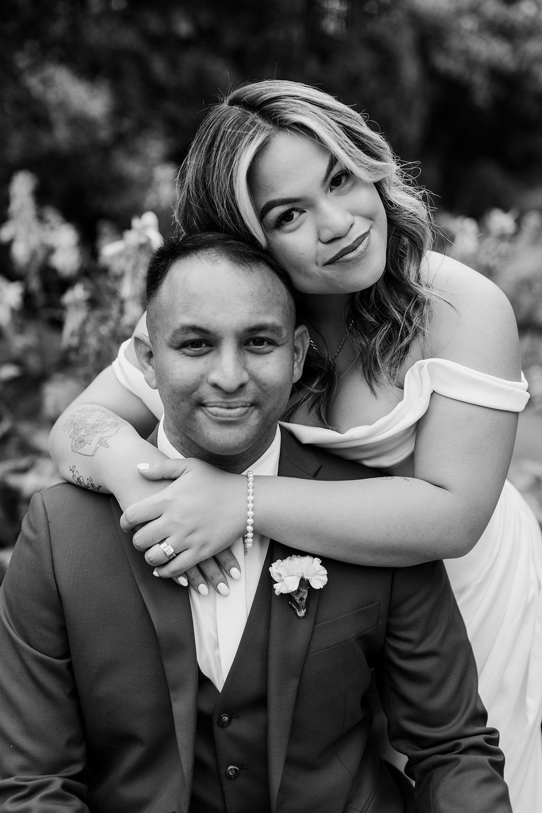 A black and white wedding portrait of a couple embracing outdoors, with the bride hugging the groom from behind.