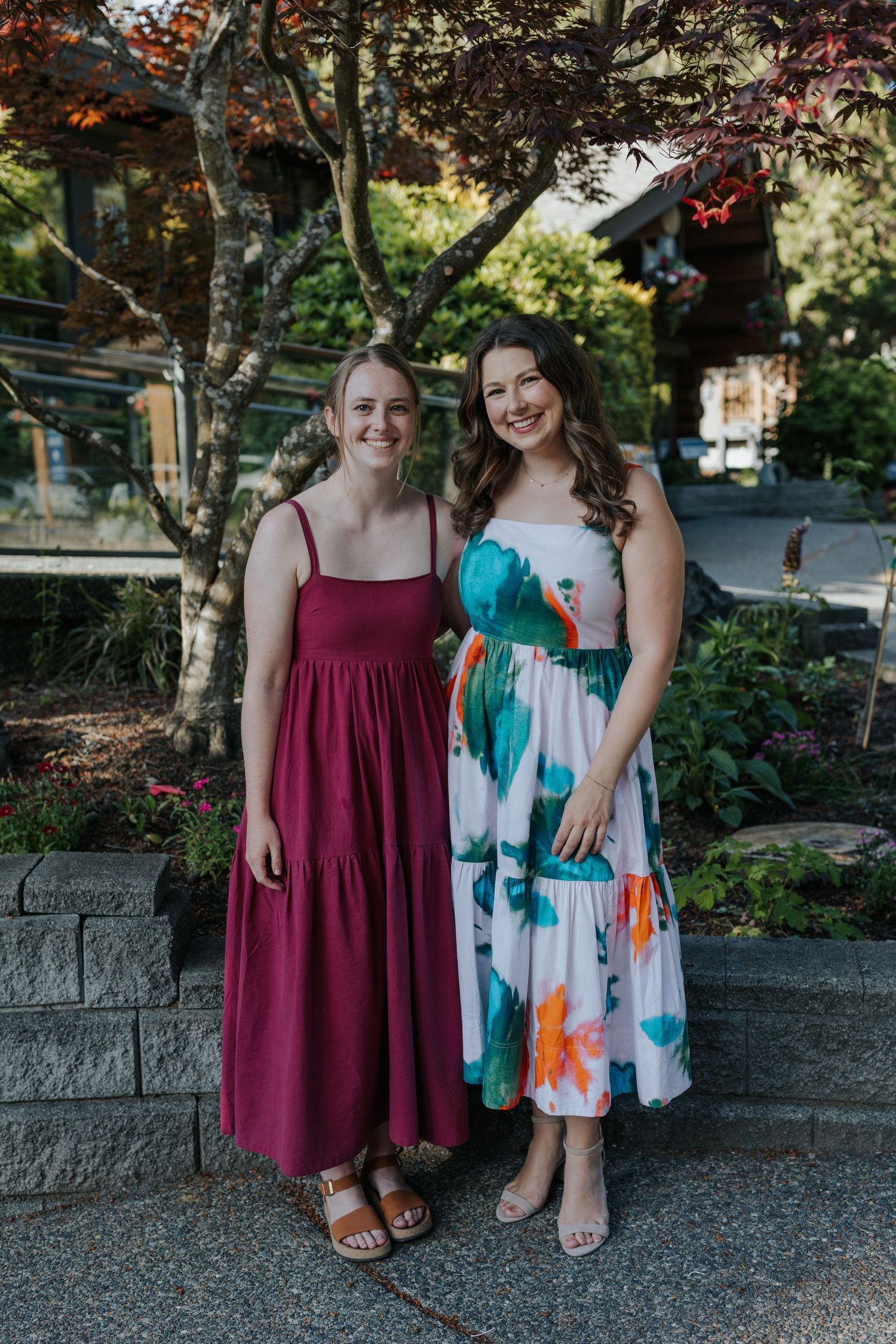 Two friends in summer dresses smile together on a stone path surrounded by greenery.