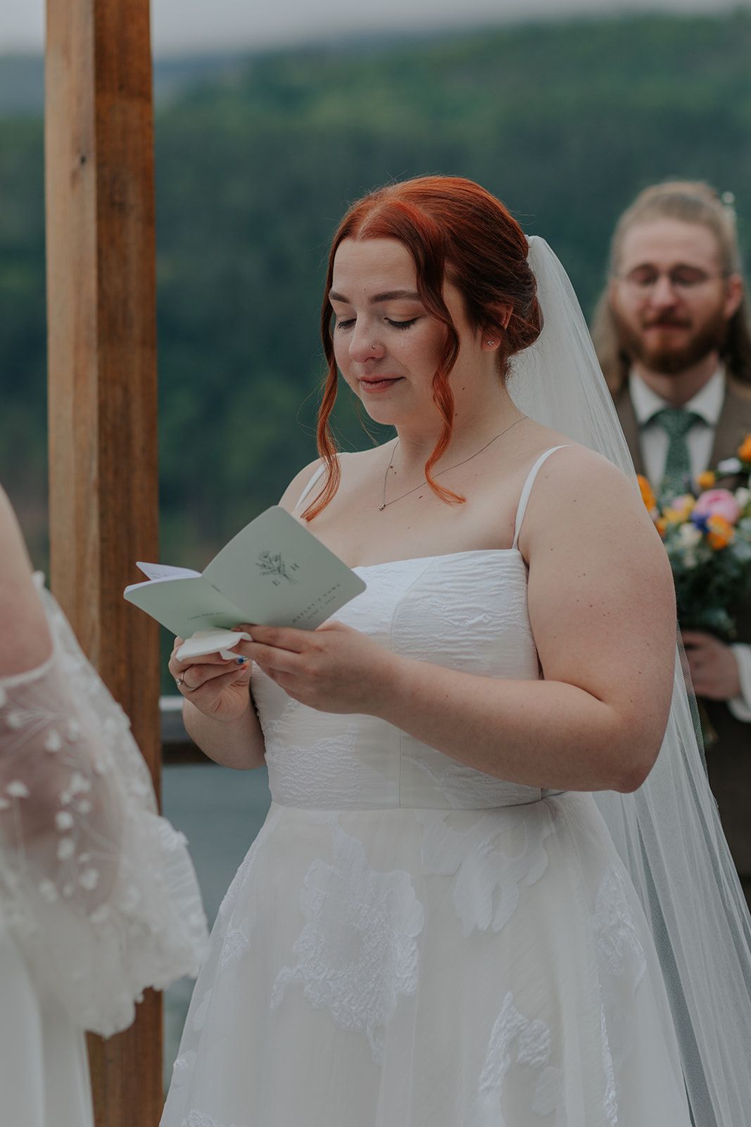 Bride in a white wedding dress reading from a paper during an outdoor ceremony.