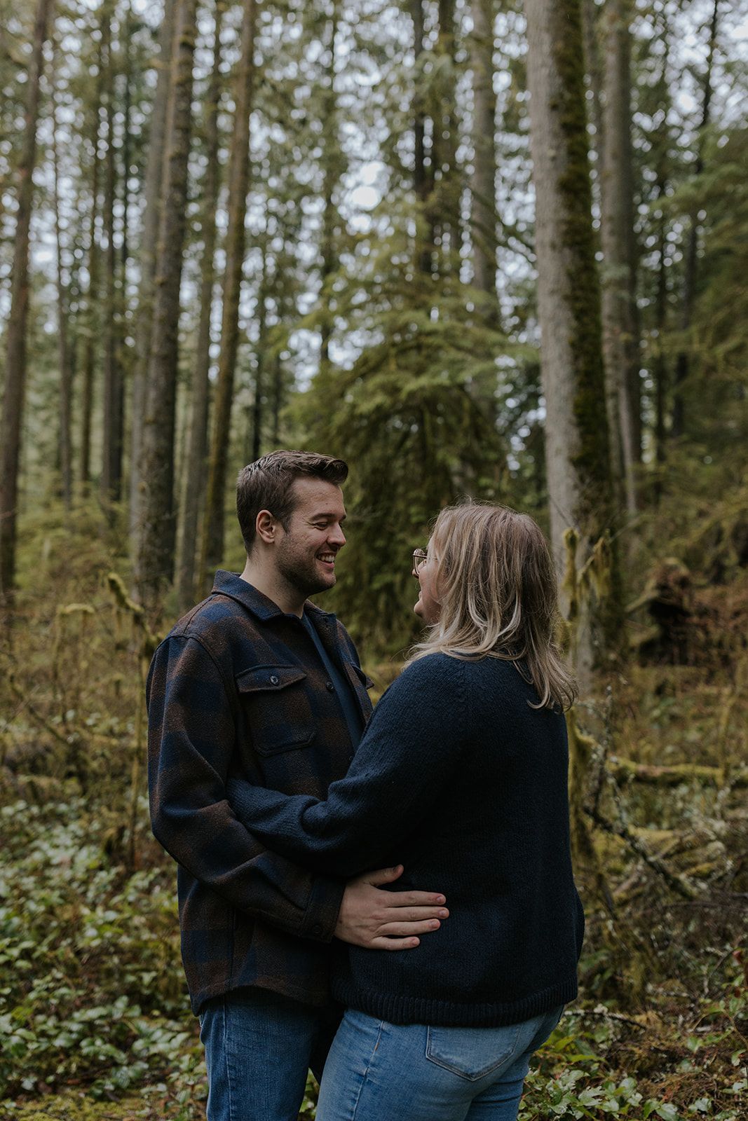 A couple embraces in a lush forest setting surrounded by tall evergreen trees.