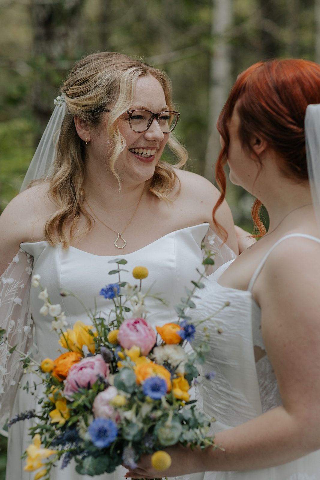 Two people in elegant white wedding dresses share joyful laughter while holding vibrant bouquets.