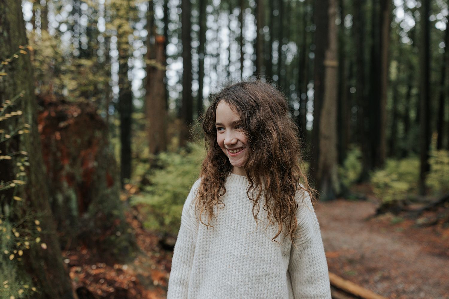 Woman in cream sweater smiling while enjoying time outdoors in a wooded area.