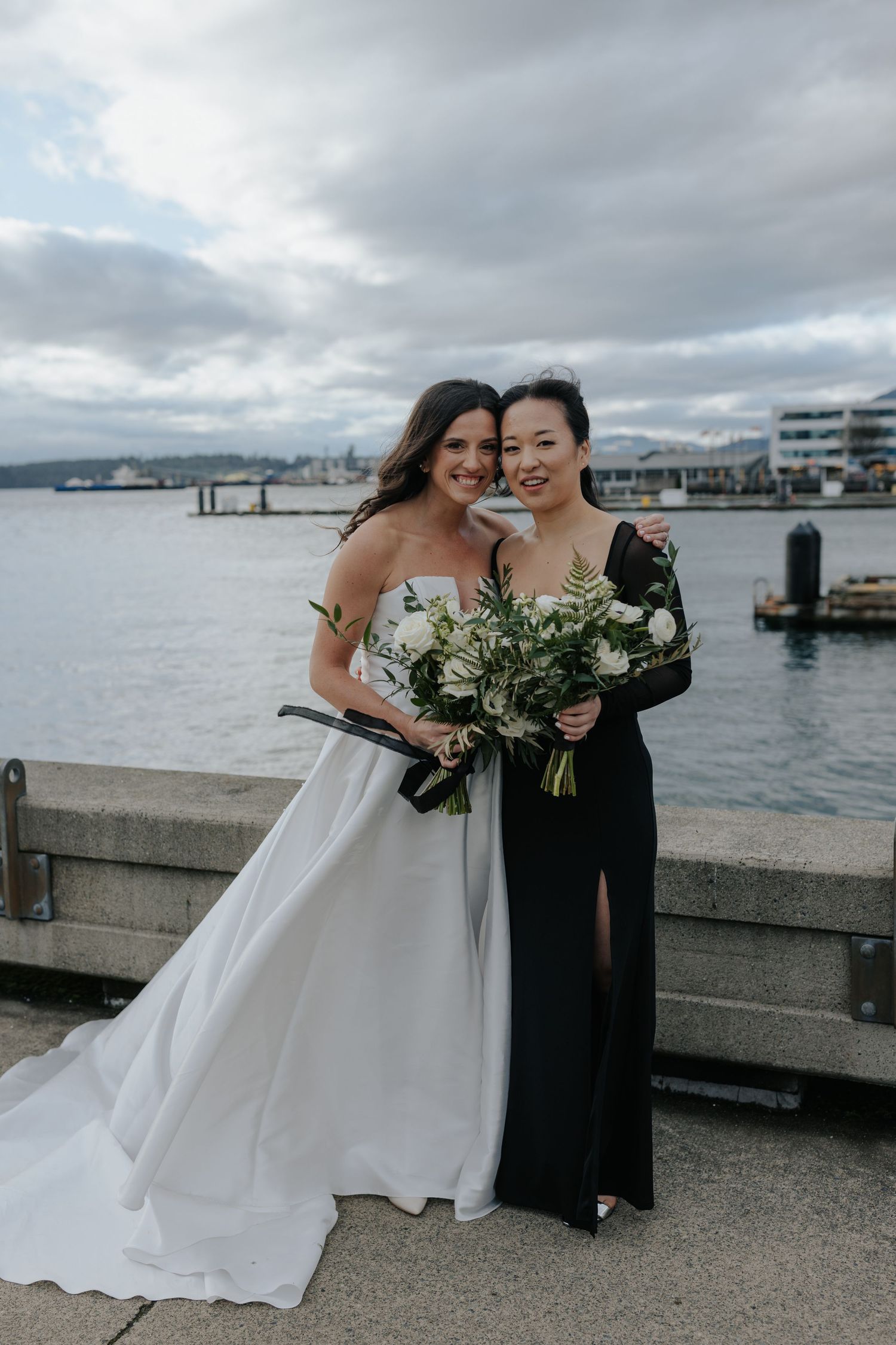 Wedding couple and attendant pose by waterfront pier with cloudy city skyline in background.