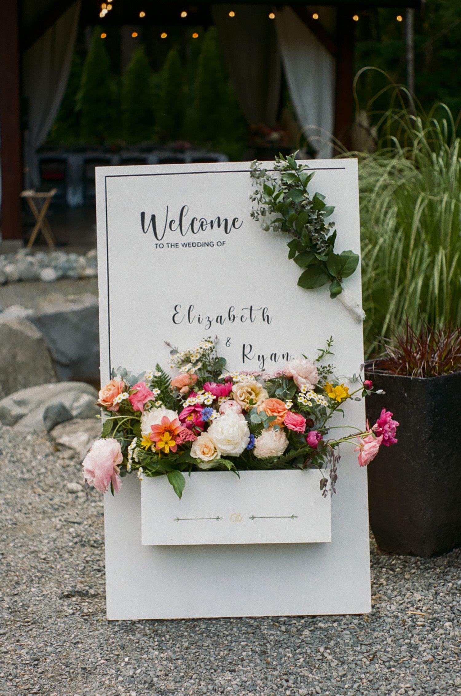 Elegant wedding welcome sign decorated with fresh flowers and greenery against a stone pathway.