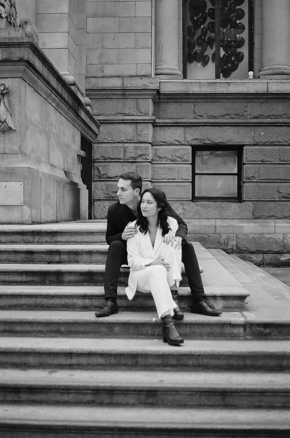 A couple sits on stone steps of a historic building in black and white.