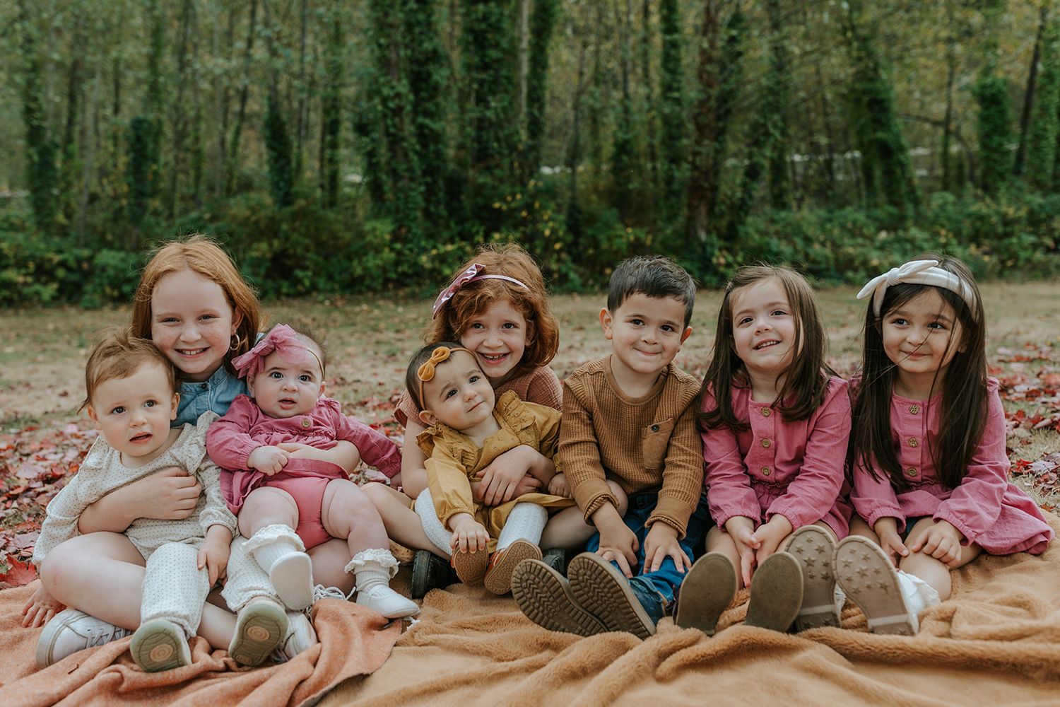 A group of children sit together on a brown blanket for an outdoor autumn portrait.