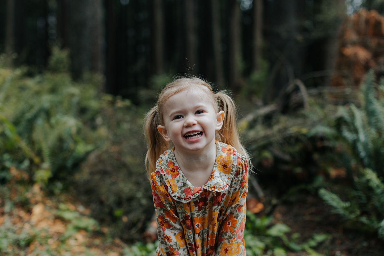 Child plays among autumn foliage wearing bright orange floral dress in forest setting.