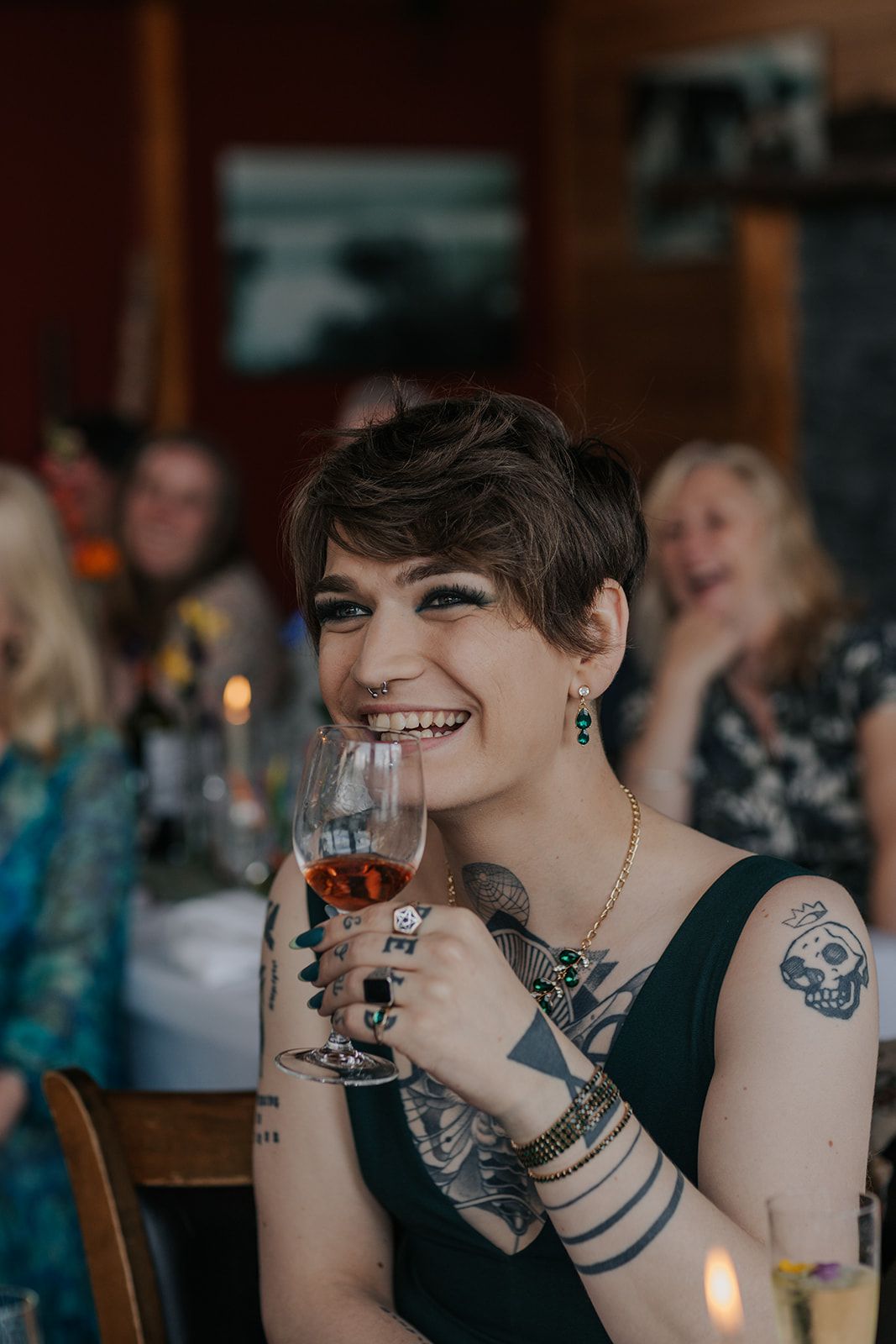 Guest with short dark hair and tattoos smiles while holding wine glass at reception.