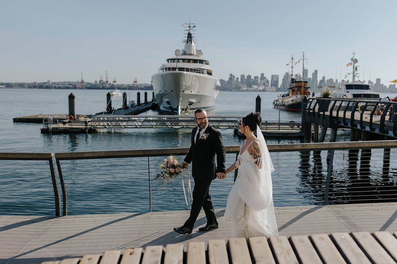 Wedding couple walking along wooden dock with city skyline in background.