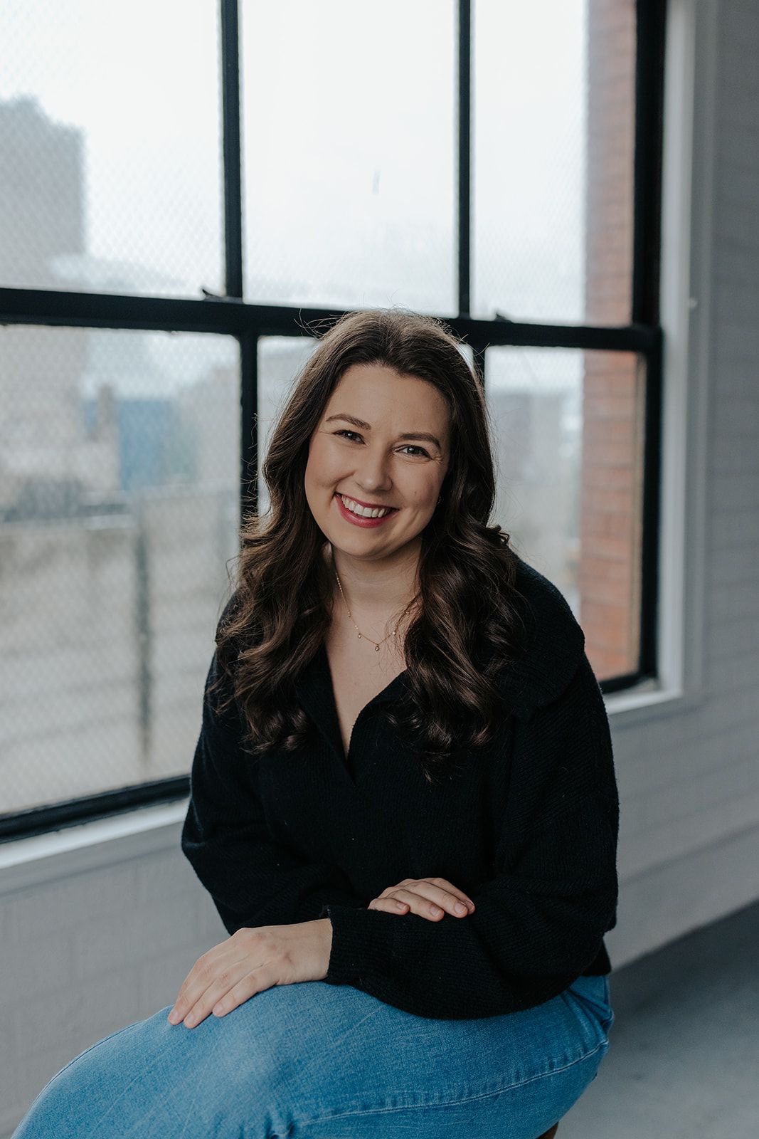 Professional headshots of woman in black top sitting by bright windows in modern office space.