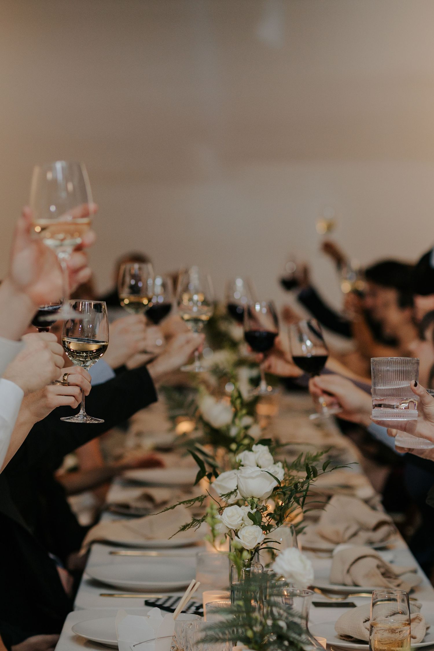 Wedding guests raise wine glasses in a toast at an elegant reception dinner table decorated with white floral centerpieces.