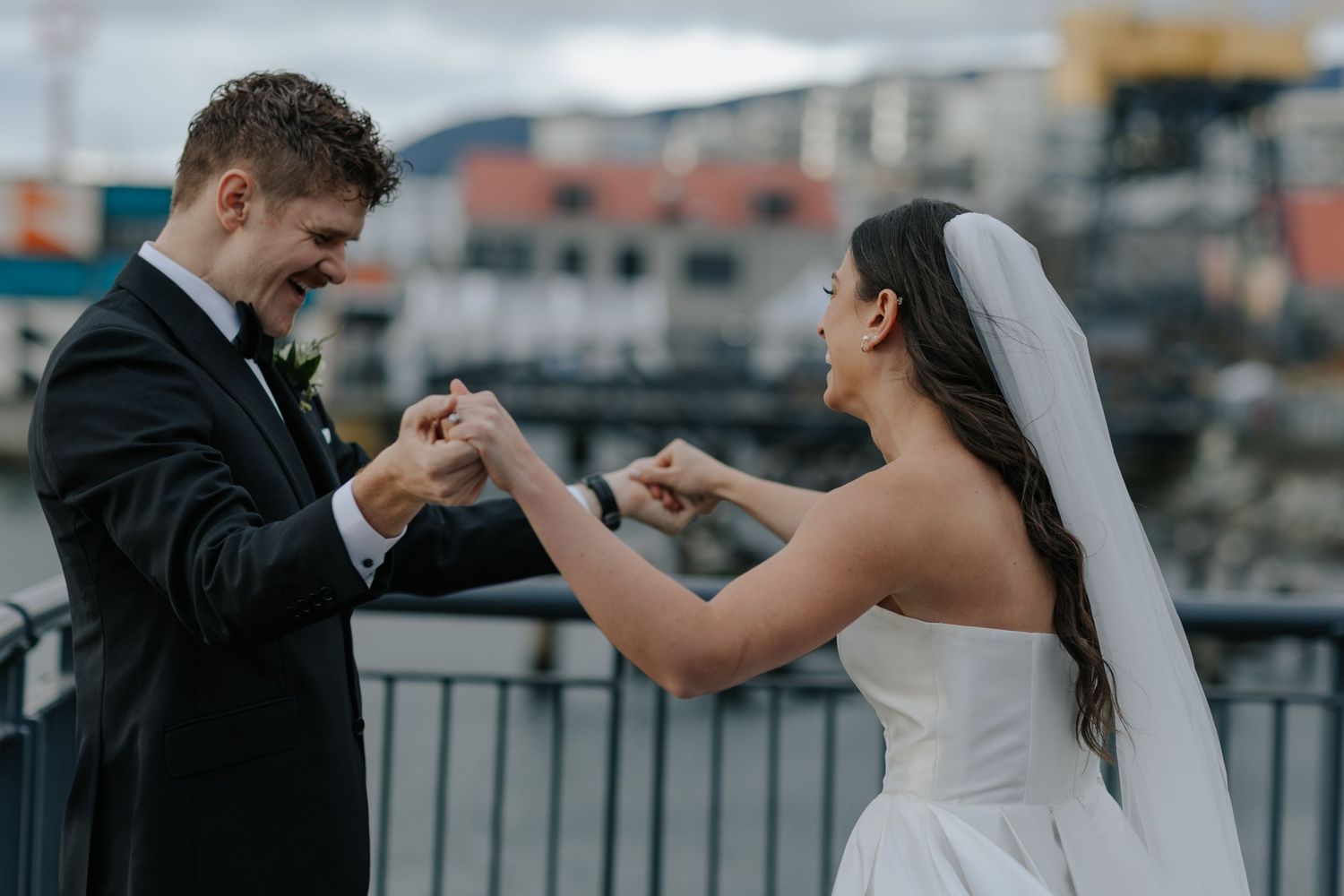 A joyful wedding couple dancing together on a waterfront boardwalk with a city skyline in the background.