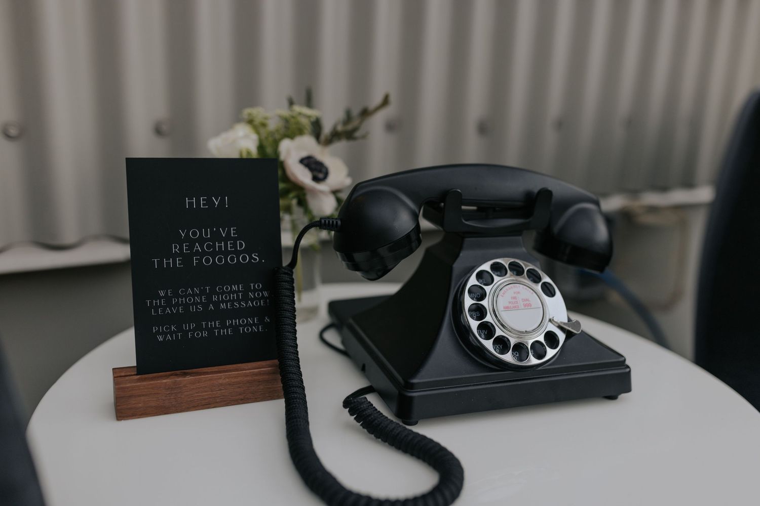 A vintage black rotary telephone sits next to a small event menu sign on a white surface.