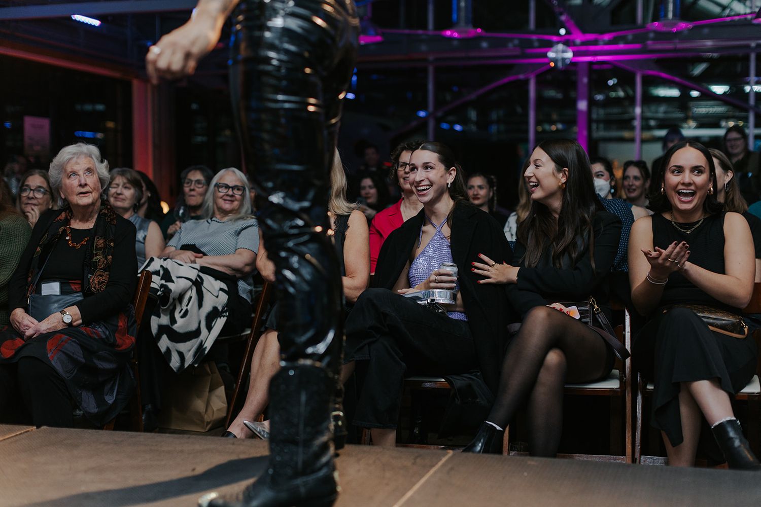 Audience members seated in rows watch a performance in a dimly lit venue with purple lighting.