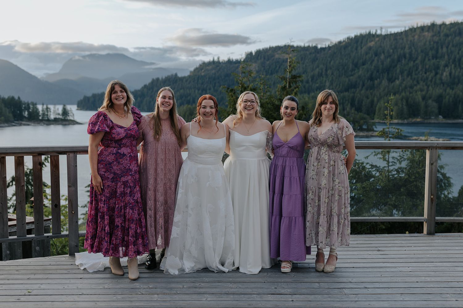 A wedding party in purple and white dresses poses on a deck overlooking a scenic mountain lake.