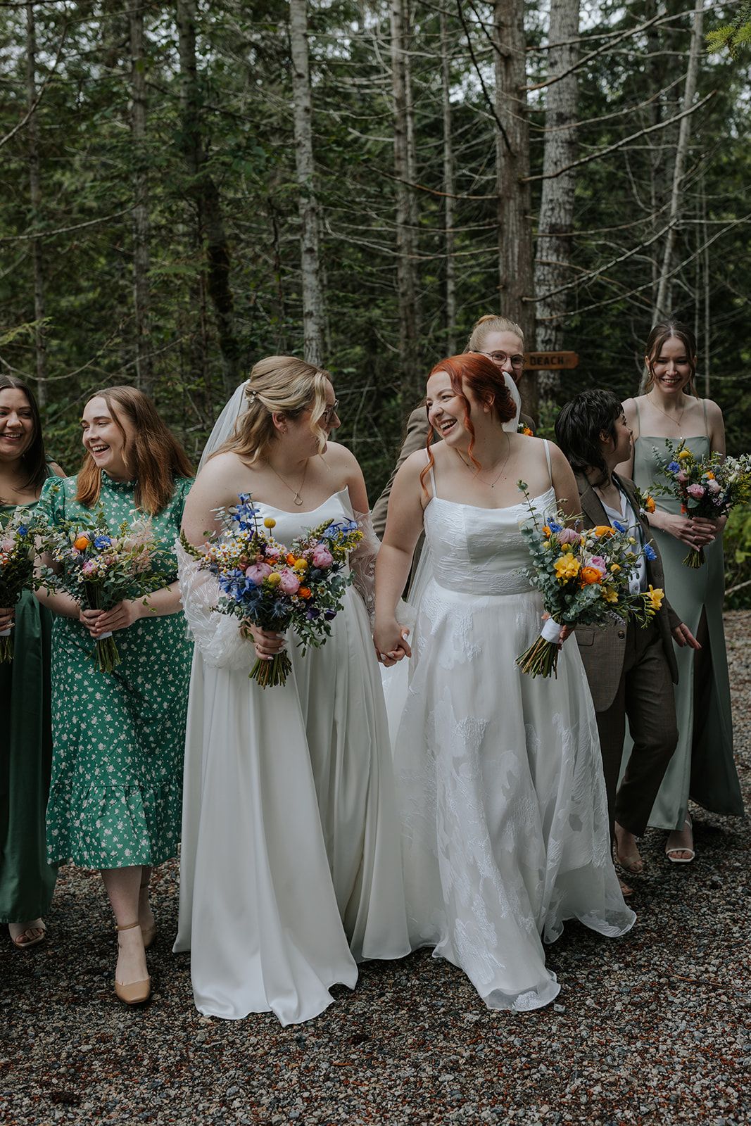 Wedding party members in white and polka dot green dresses hold colorful bouquets in forest.