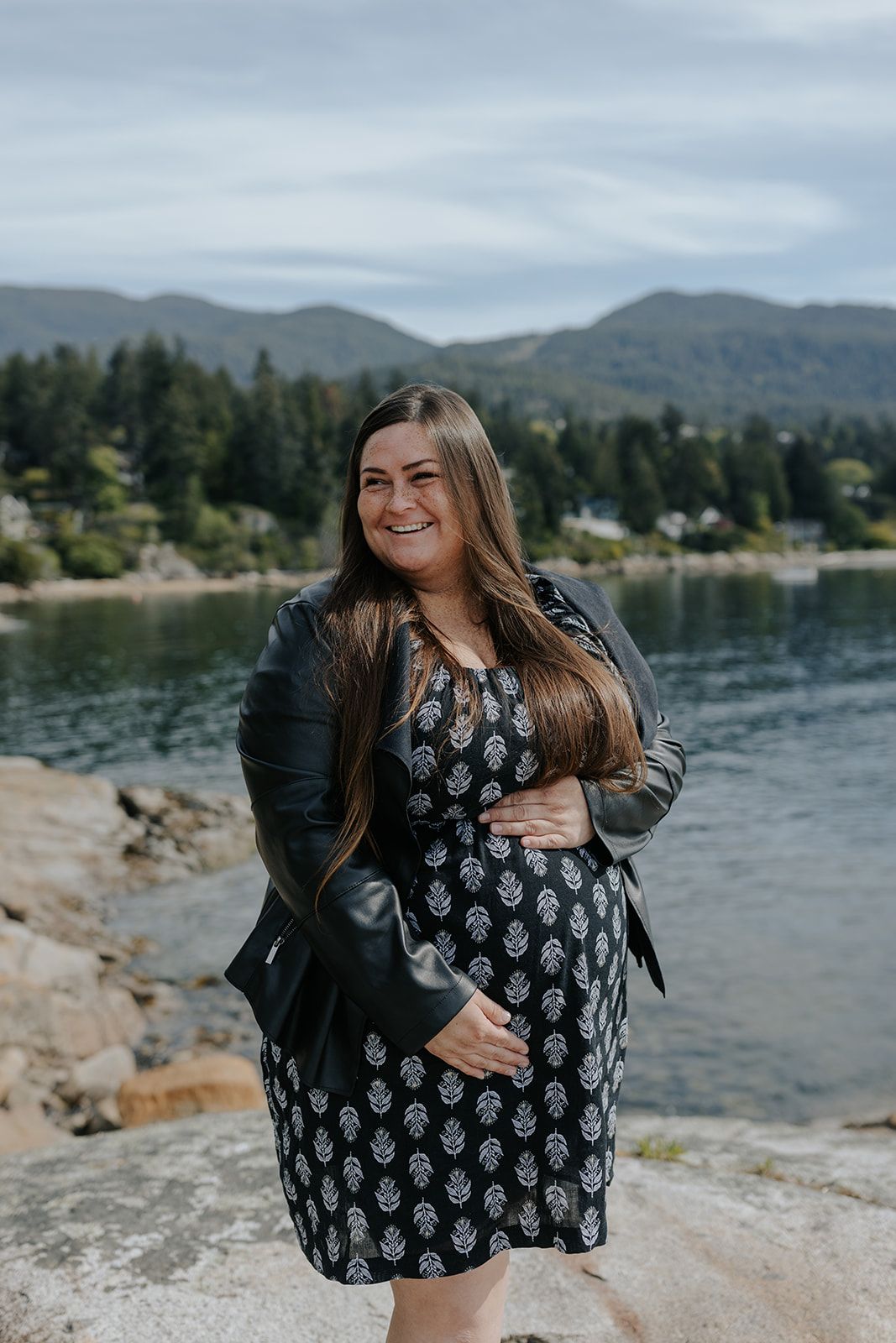 Woman in floral dress laughs joyfully on rocky coastline with mountains and lake in background.