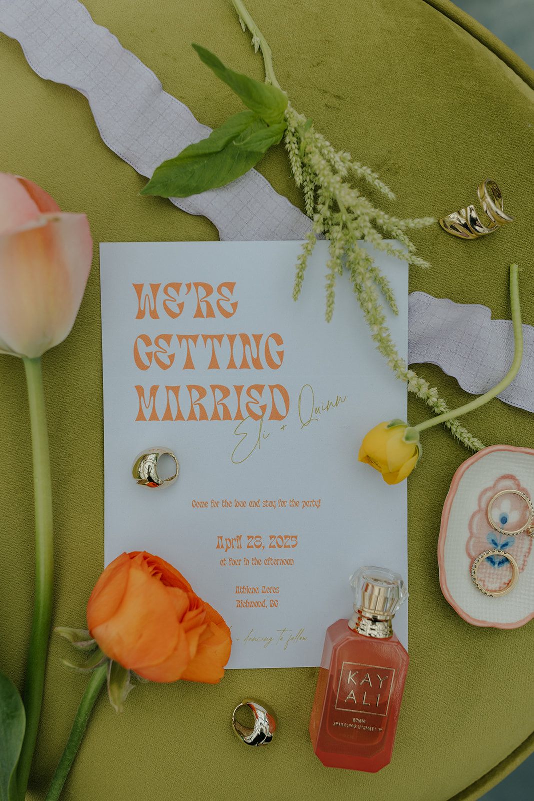 Wedding invitation flatlay with rings, perfume bottle, and peach roses on green background.