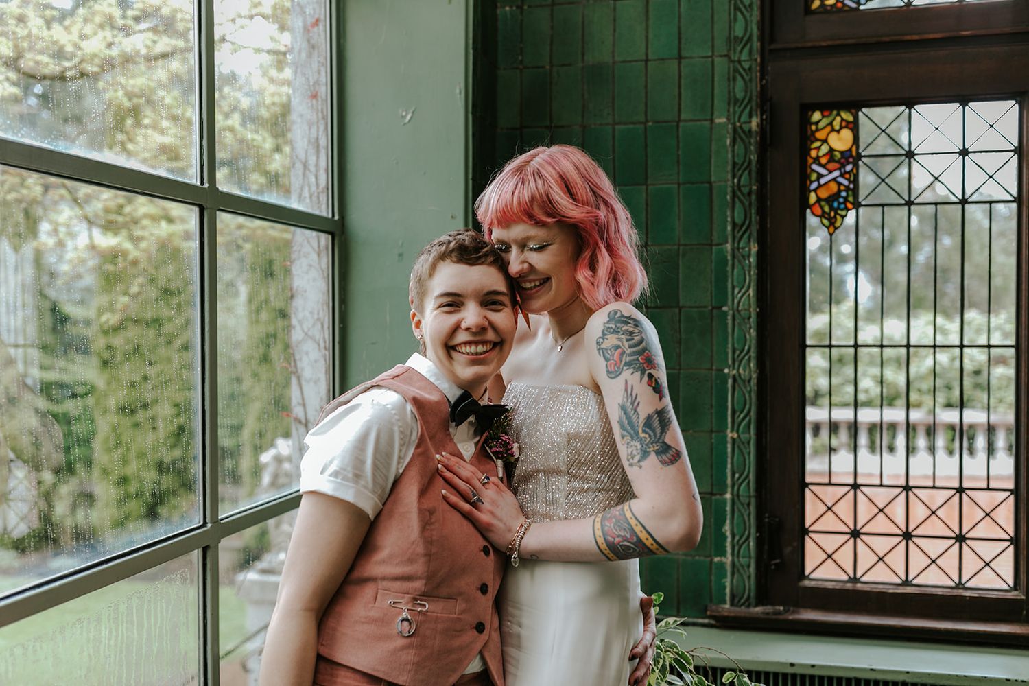 A couple in wedding attire share a joyful moment together near a green-framed window.