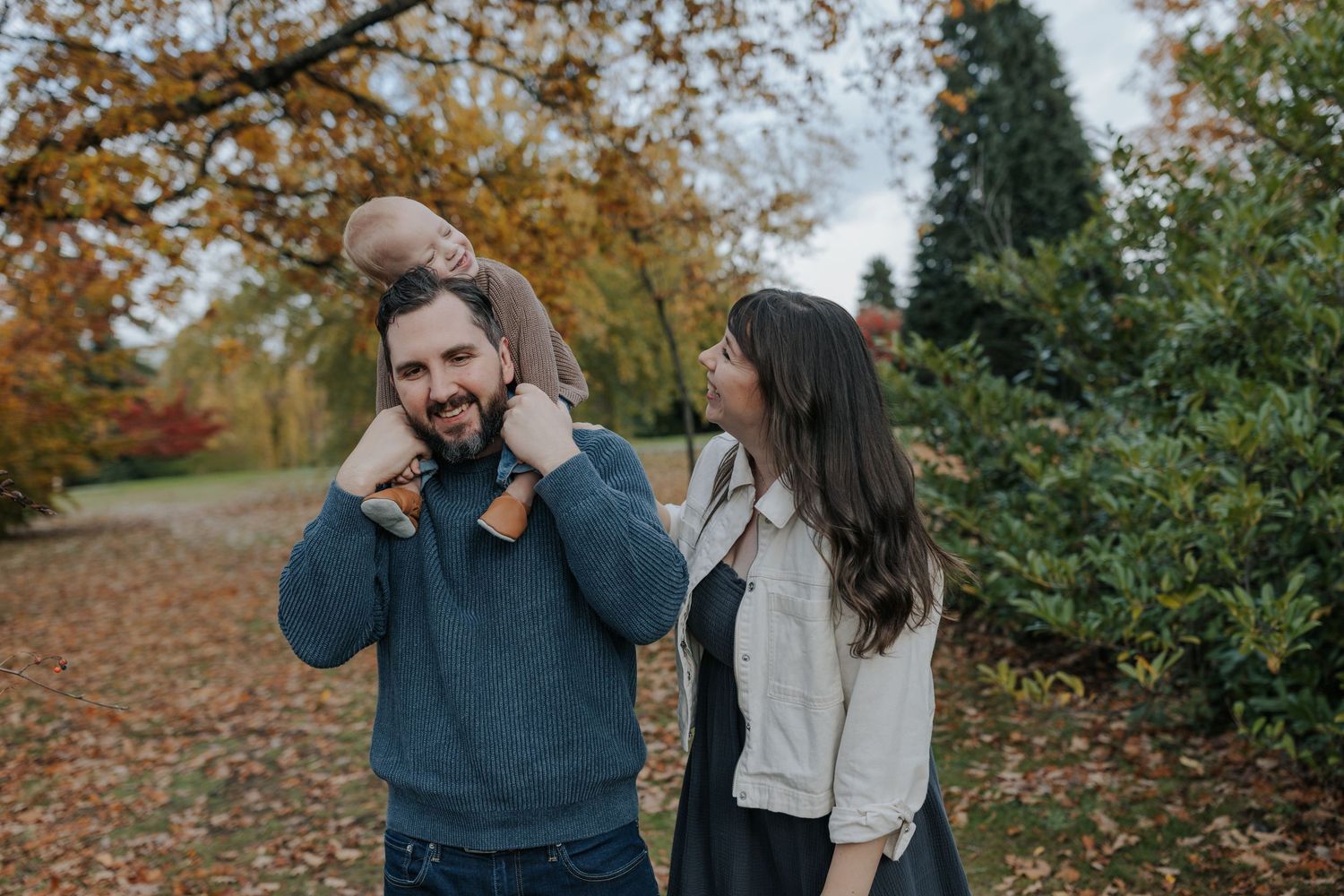 Family enjoying autumn park walk, father carrying baby on shoulders while mother smiles beside them.