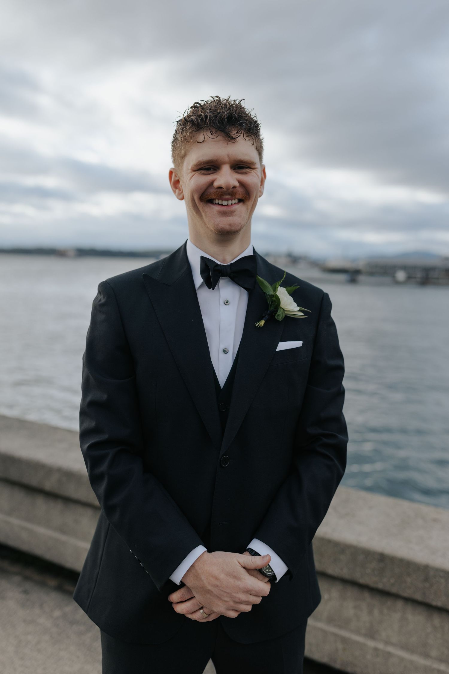 A groom in classic black tuxedo stands by the waterfront on his wedding day.
