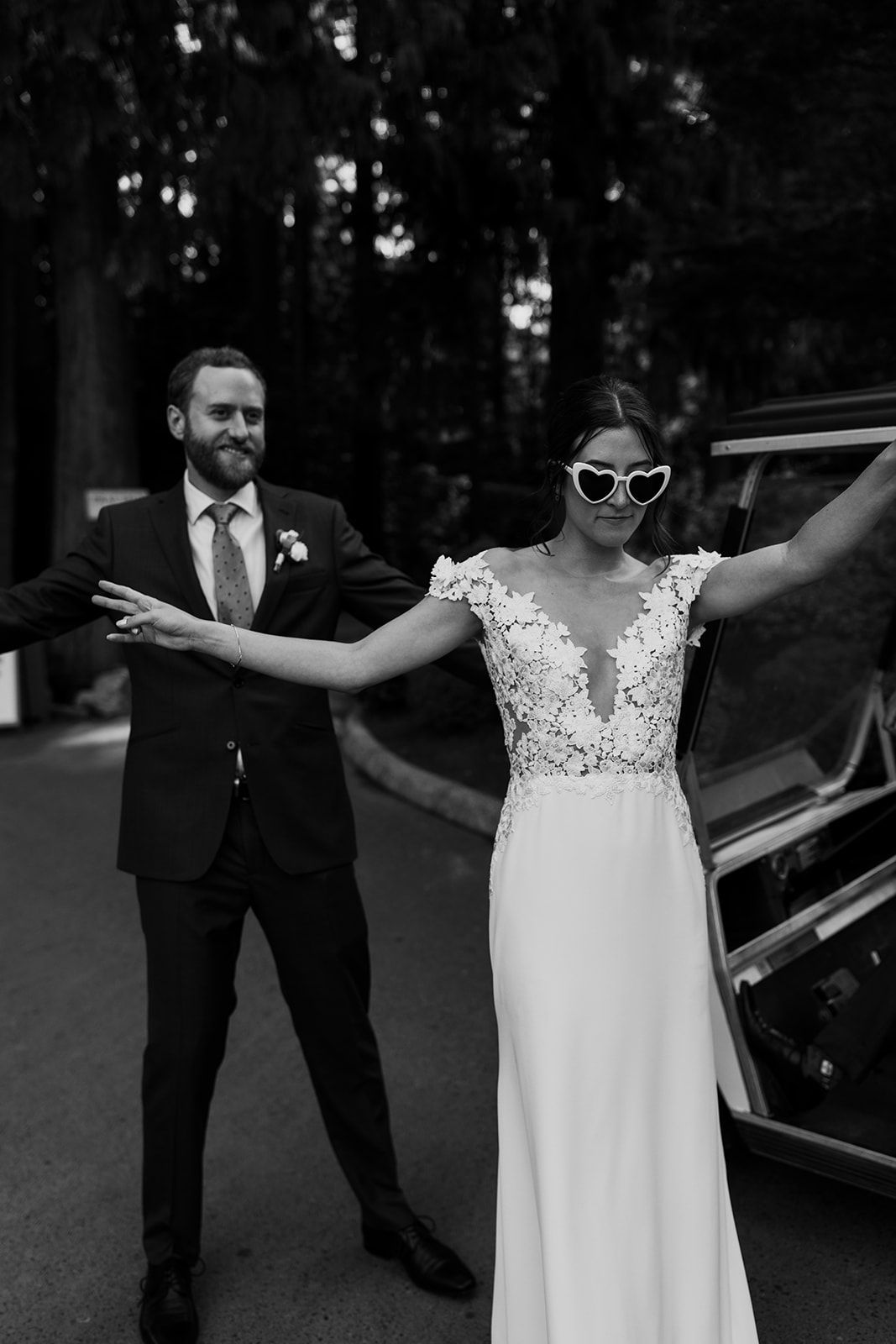 A couple in wedding attire poses playfully on a forest road.