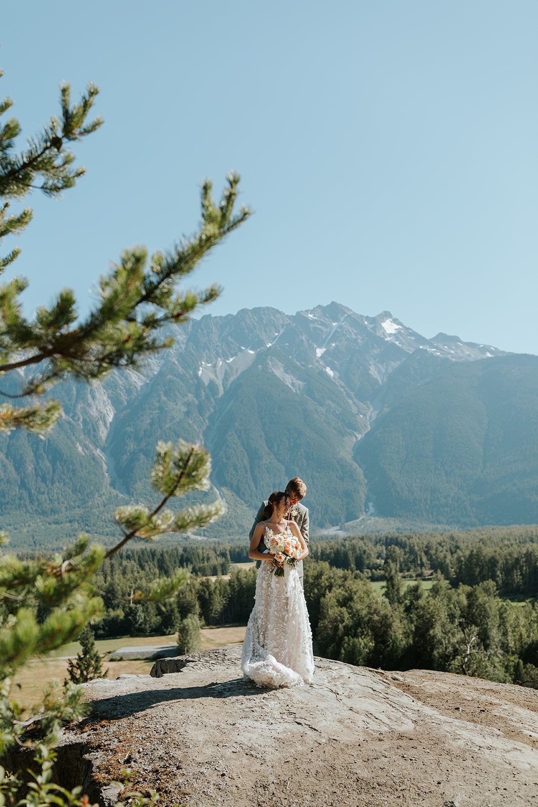 A couple embraces on a rocky outcrop with snow-capped mountains in the background.