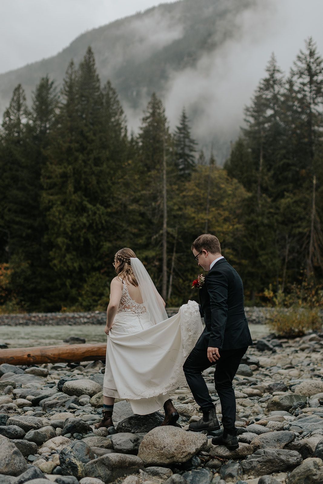 Couple walks carefully across rocky riverbed with misty mountains in background.